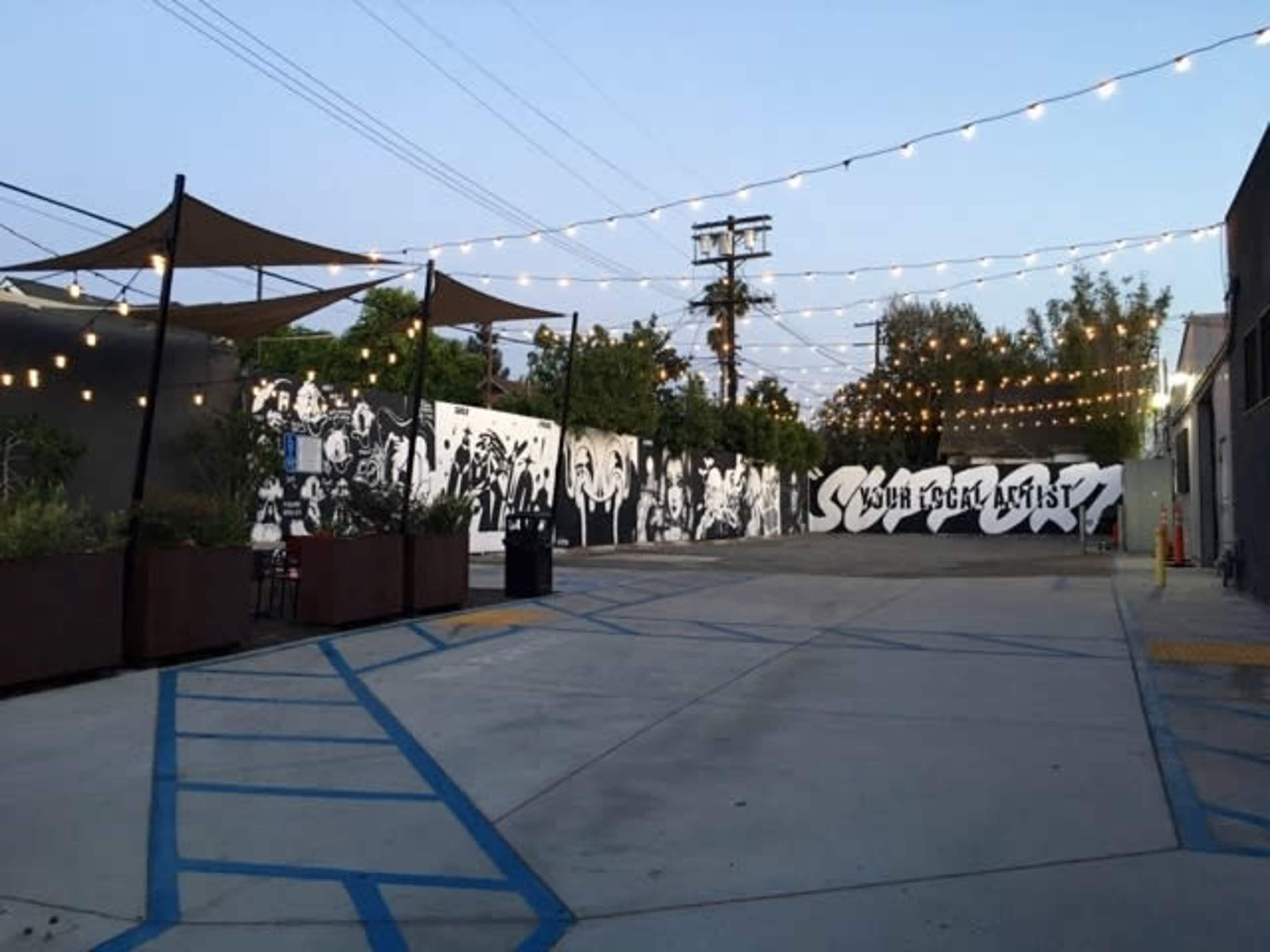 A street with a large black-and-white mural on a wall, surrounded by decorative string lights and shaded seating areas.