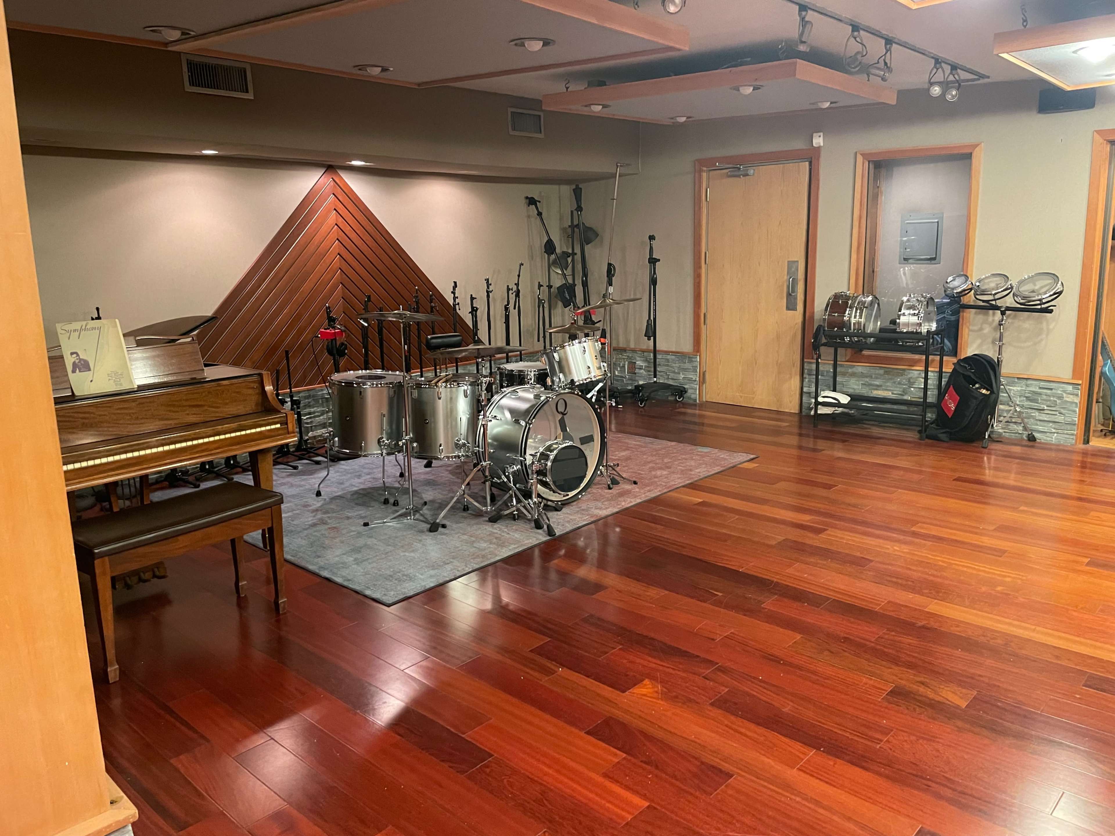 A music studio with a grand piano on one side and a drum set on a rug in the center, surrounded by microphones and various instruments on the walls.