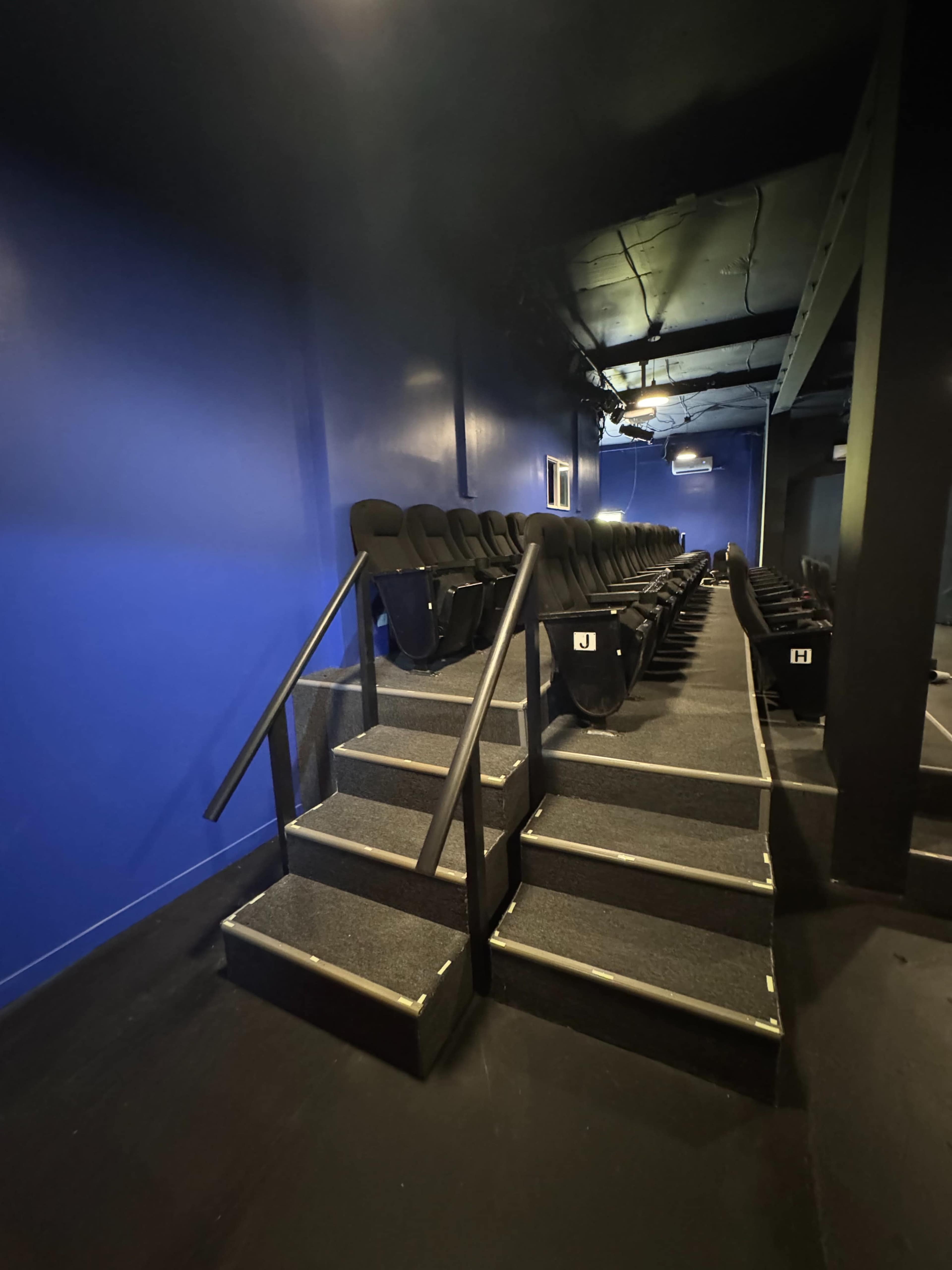 The image shows a row of theater seats arranged on a stepped platform in a dimly lit space with blue walls.