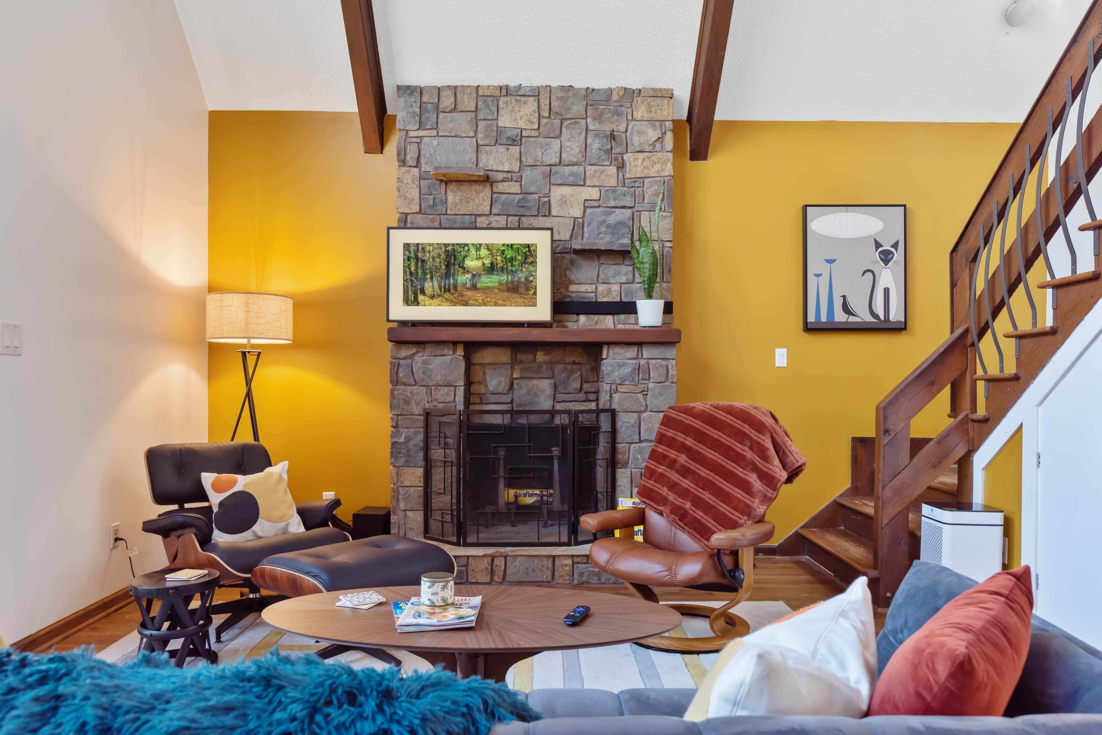 A cozy living room with a stone fireplace, a yellow accent wall, and a mix of modern and eclectic furniture arranged around a coffee table.