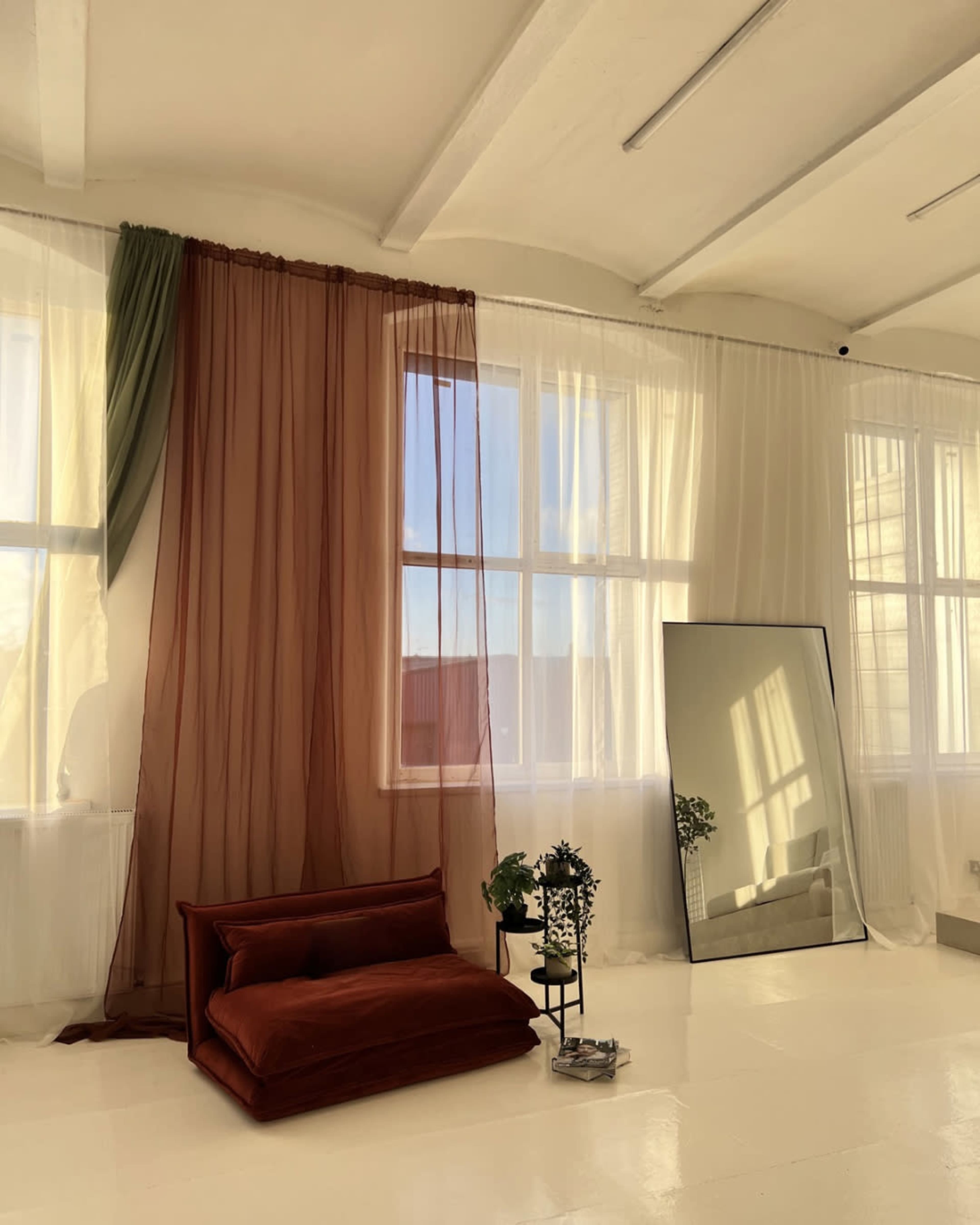 The image shows a bright room with large windows featuring sheer curtains, a brown velvet sofa, and a tall mirror beside a plant.