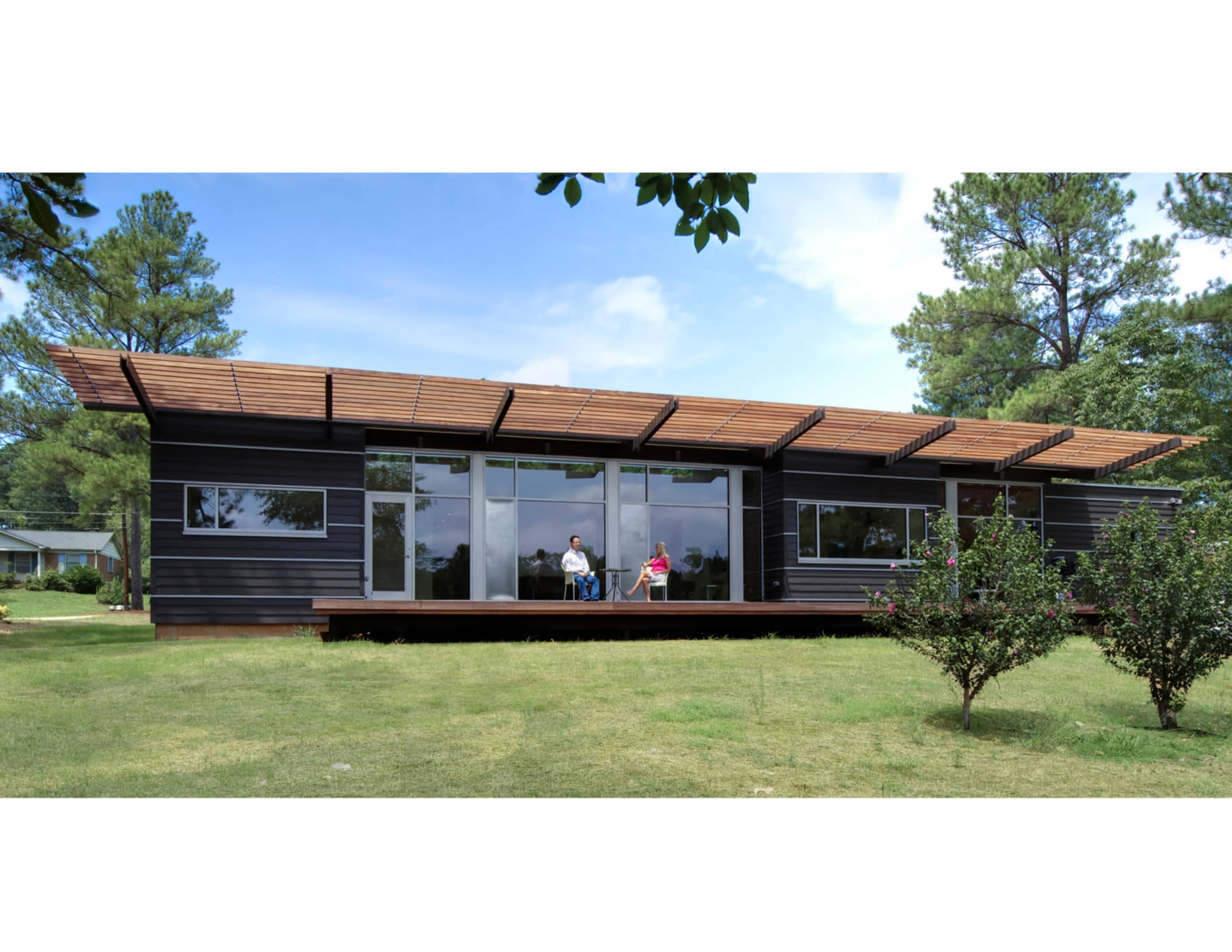 A modern house with large windows and a wooden deck is situated in a grassy area with two people seated outside, surrounded by trees.