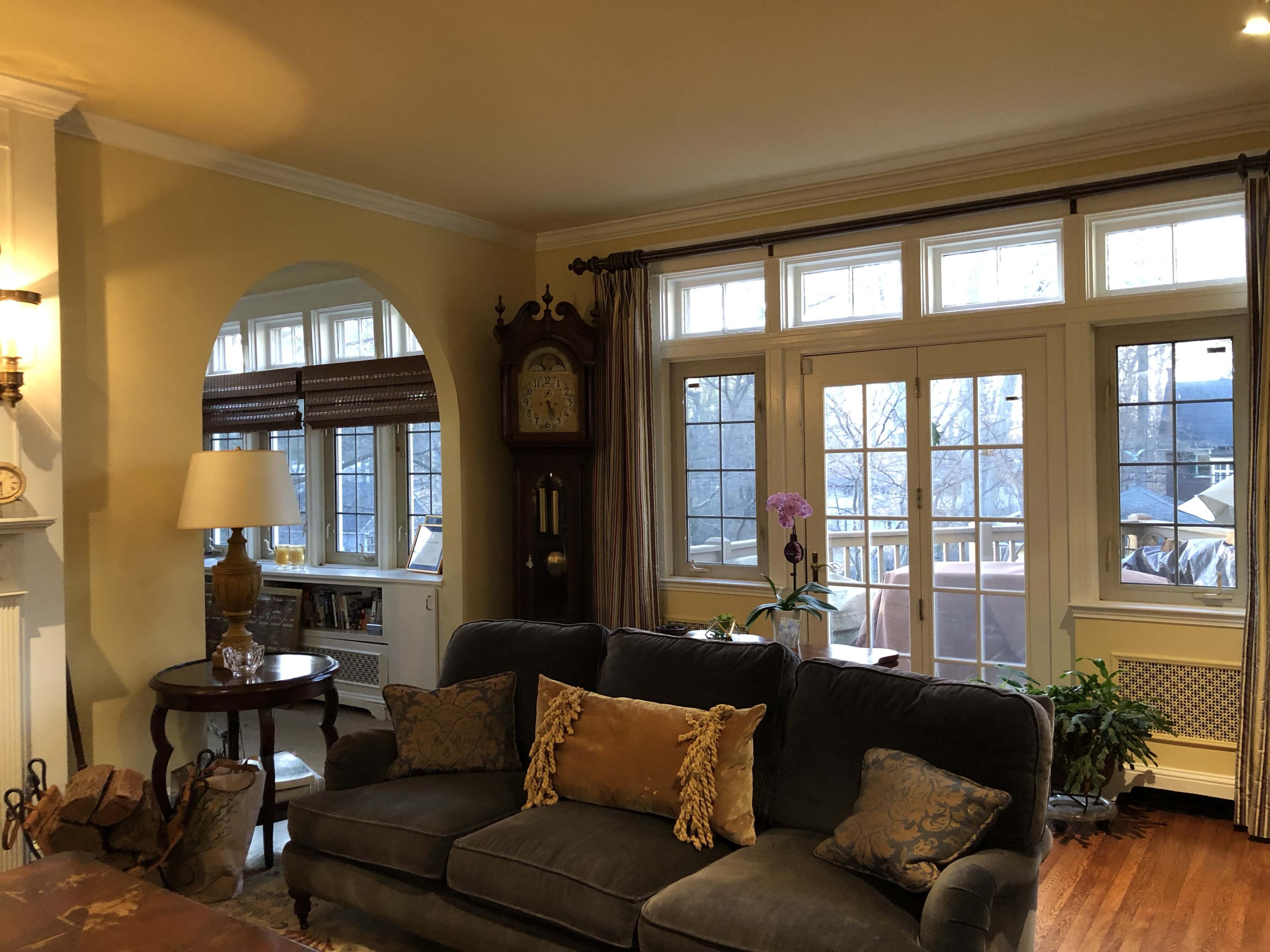 The image shows a cozy living room featuring a dark sofa, a round table with a lamp, a large clock, and large windows with light curtains.