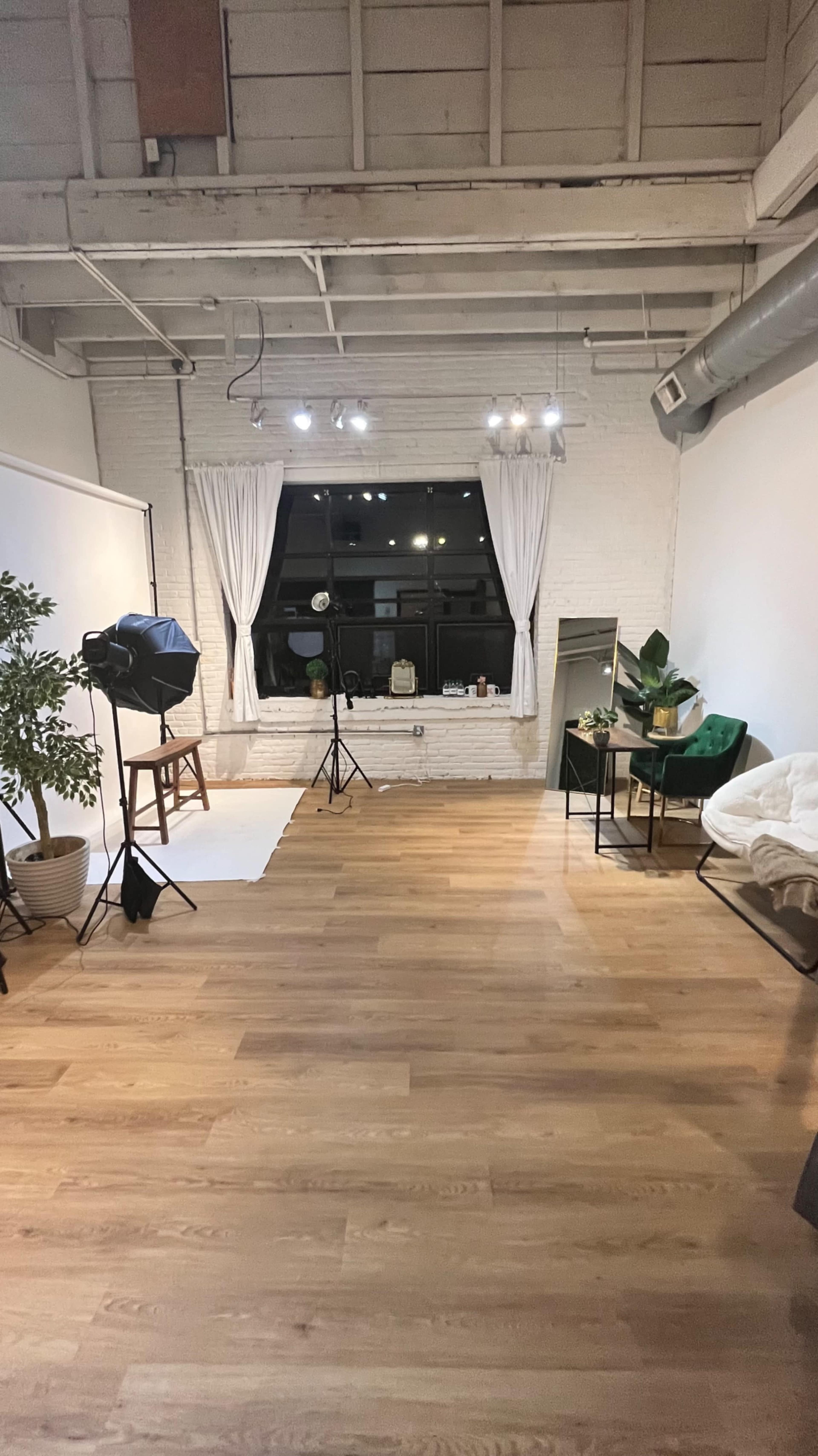 The image shows a spacious, well-lit room with wooden flooring, a seating area with green chairs, a backdrop setup for photography, and large windows dressed with white curtains.