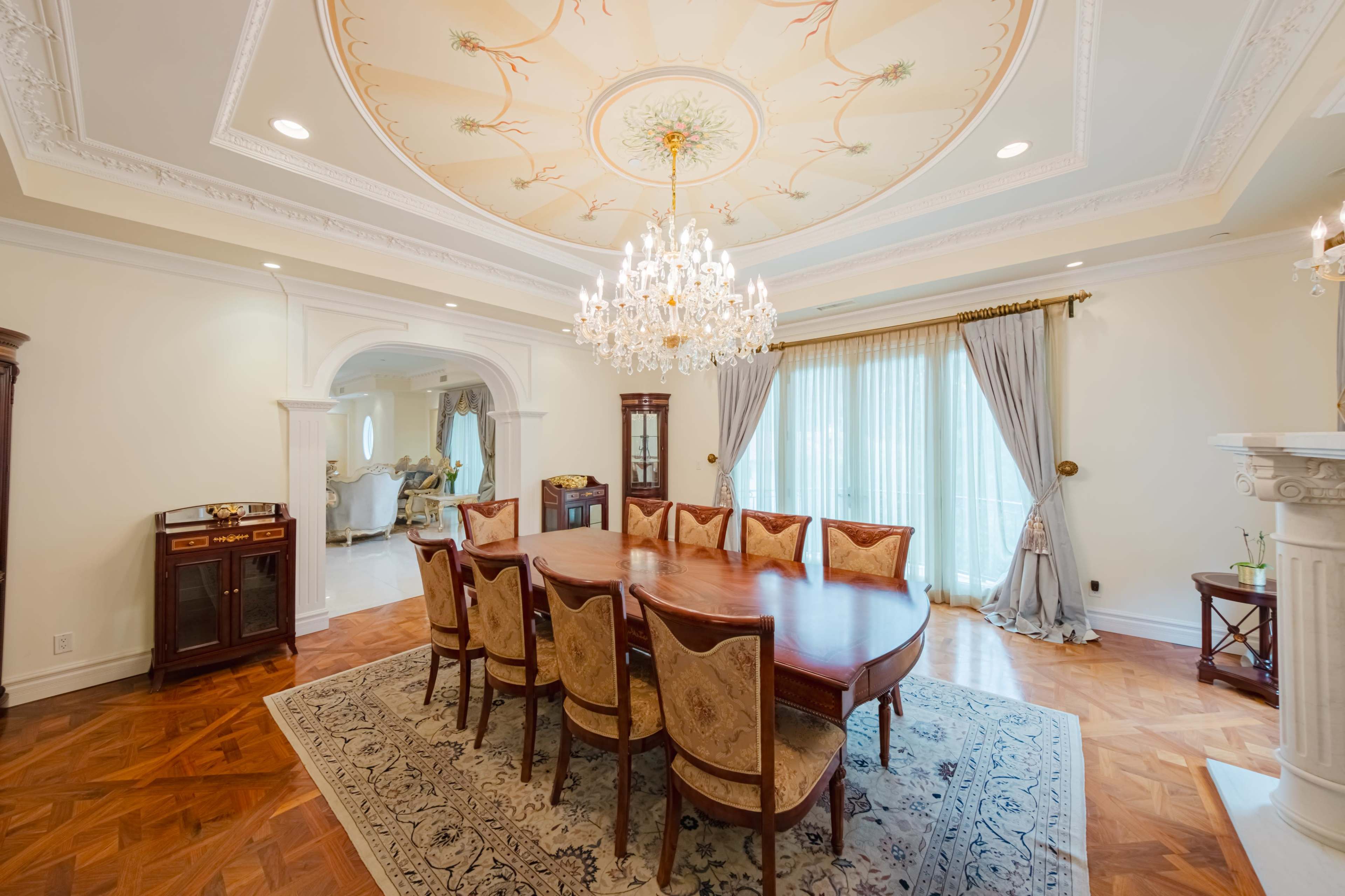 A spacious dining room features an ornate ceiling, a large wooden table surrounded by upholstered chairs, and a chandelier, with large windows draped in light curtains.