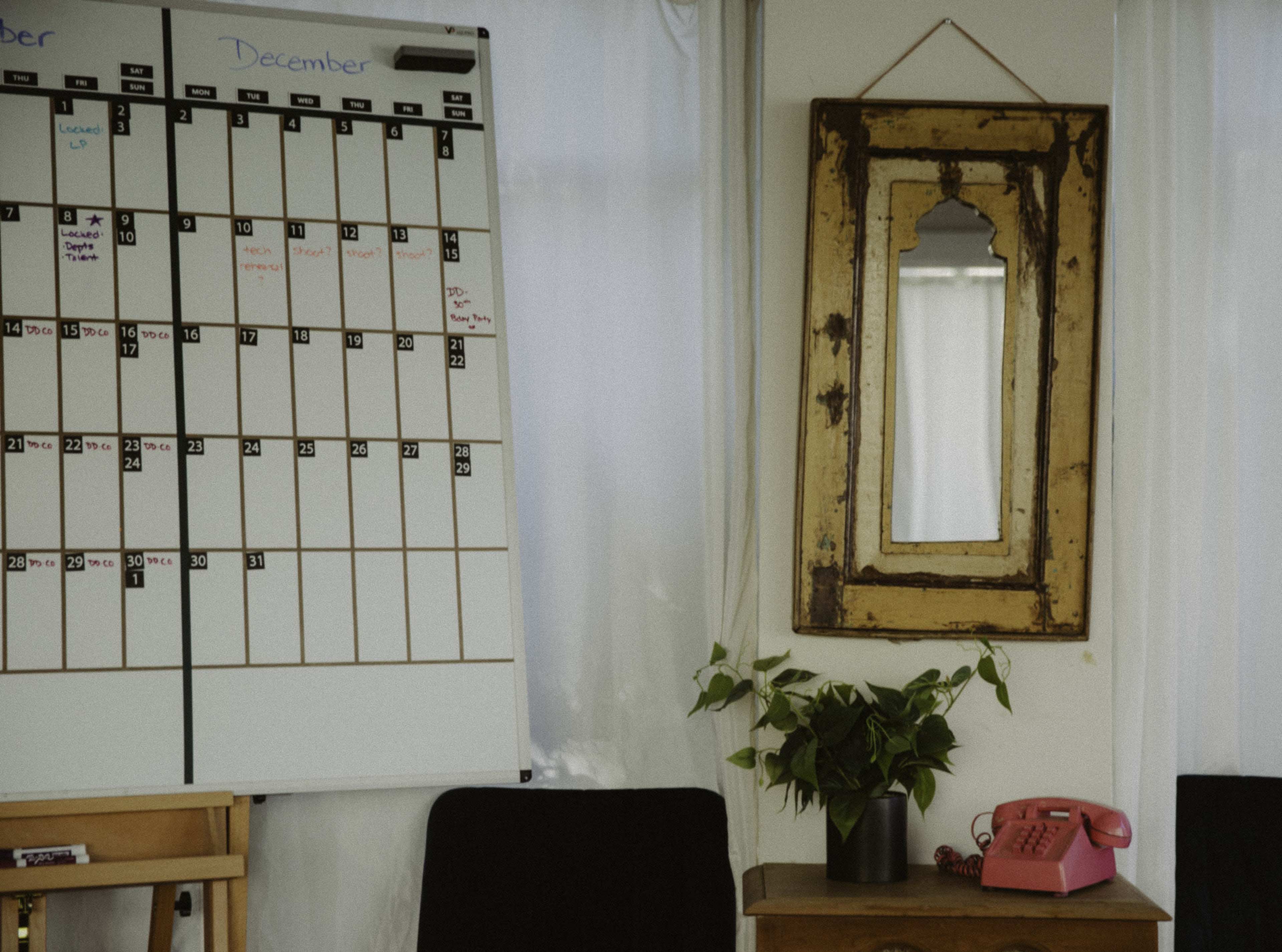 A large calendar board is mounted on the wall next to a vintage mirror, with a pink telephone and a potted plant on a wooden table.