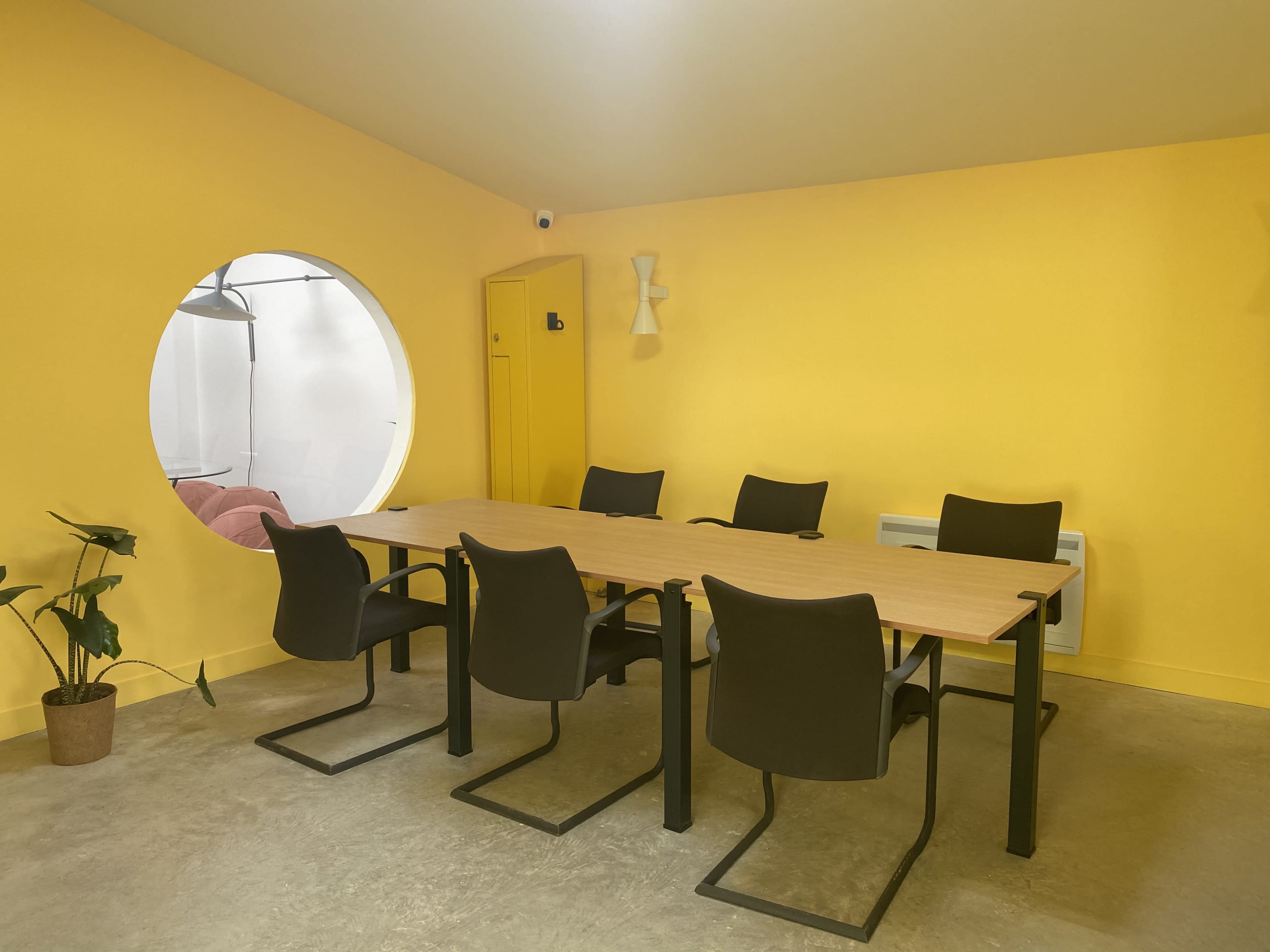 A meeting room with a large yellow wall, a wooden table surrounded by six black chairs, and a circular window leading to another room.
