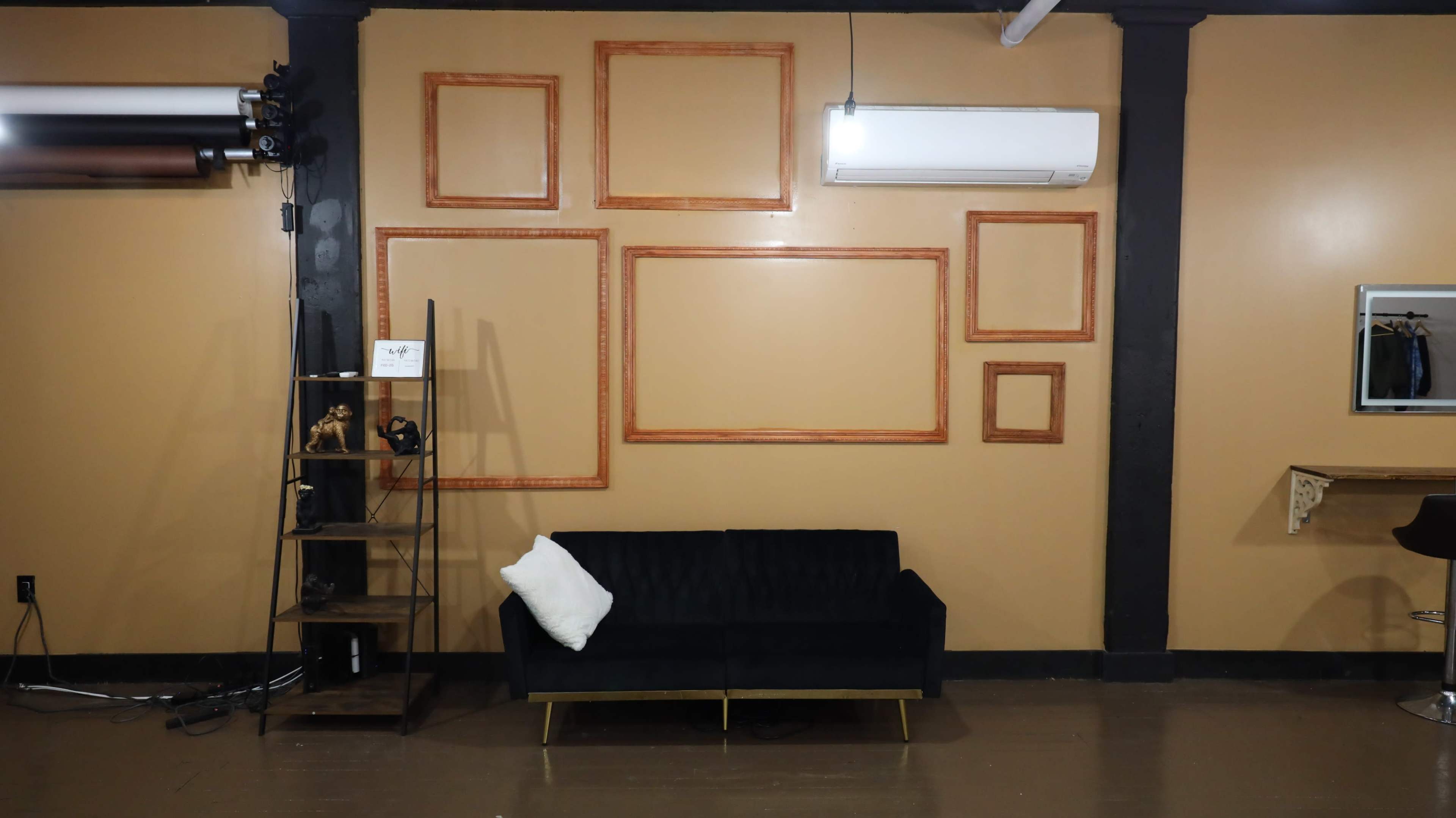 A room with a brown wall featuring several empty wooden picture frames arranged in different sizes above a black sofa with a white pillow, alongside a metallic shelf with decorative items.
