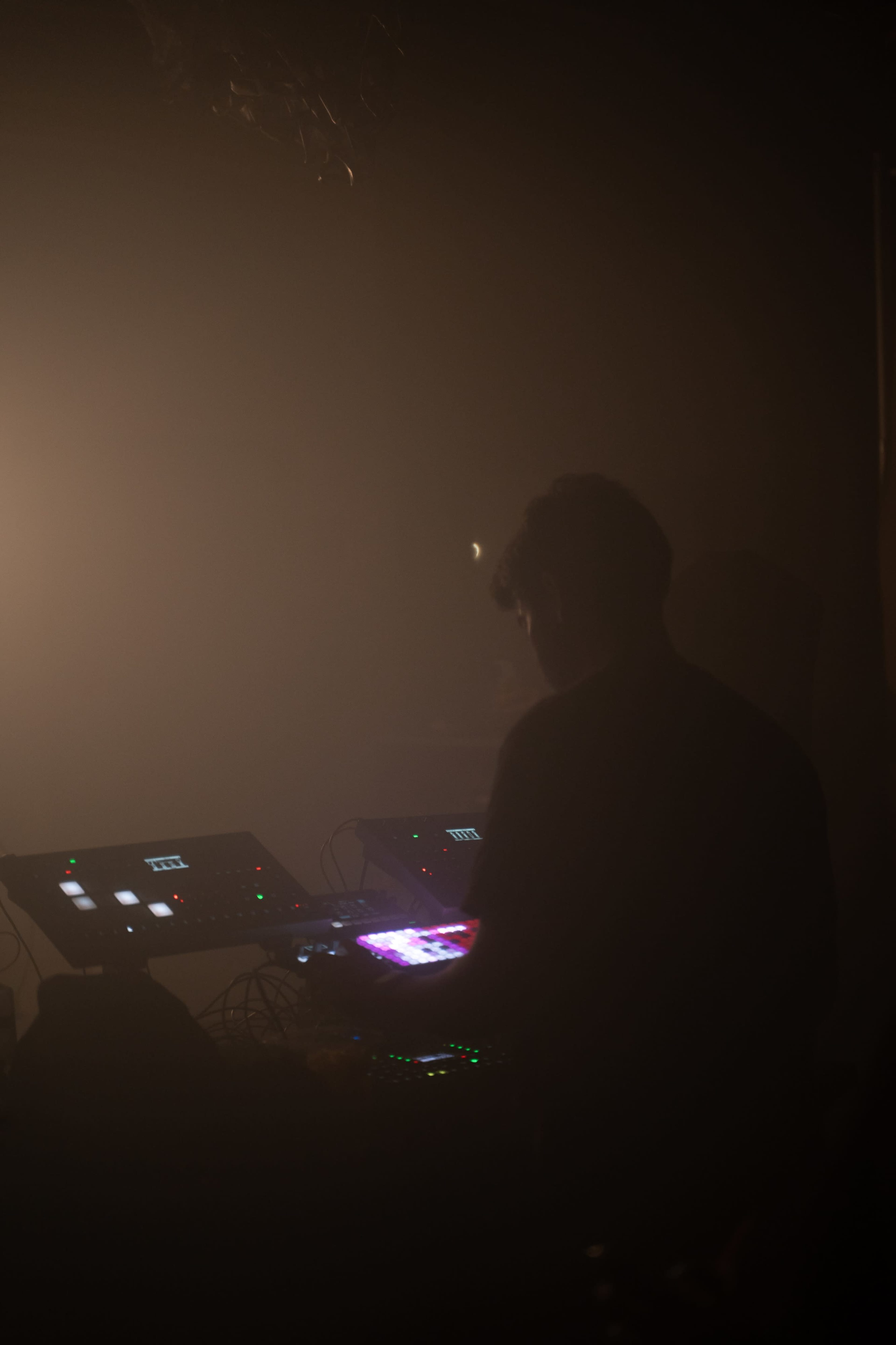 A person is operating electronic music equipment in a dimly lit, foggy environment.