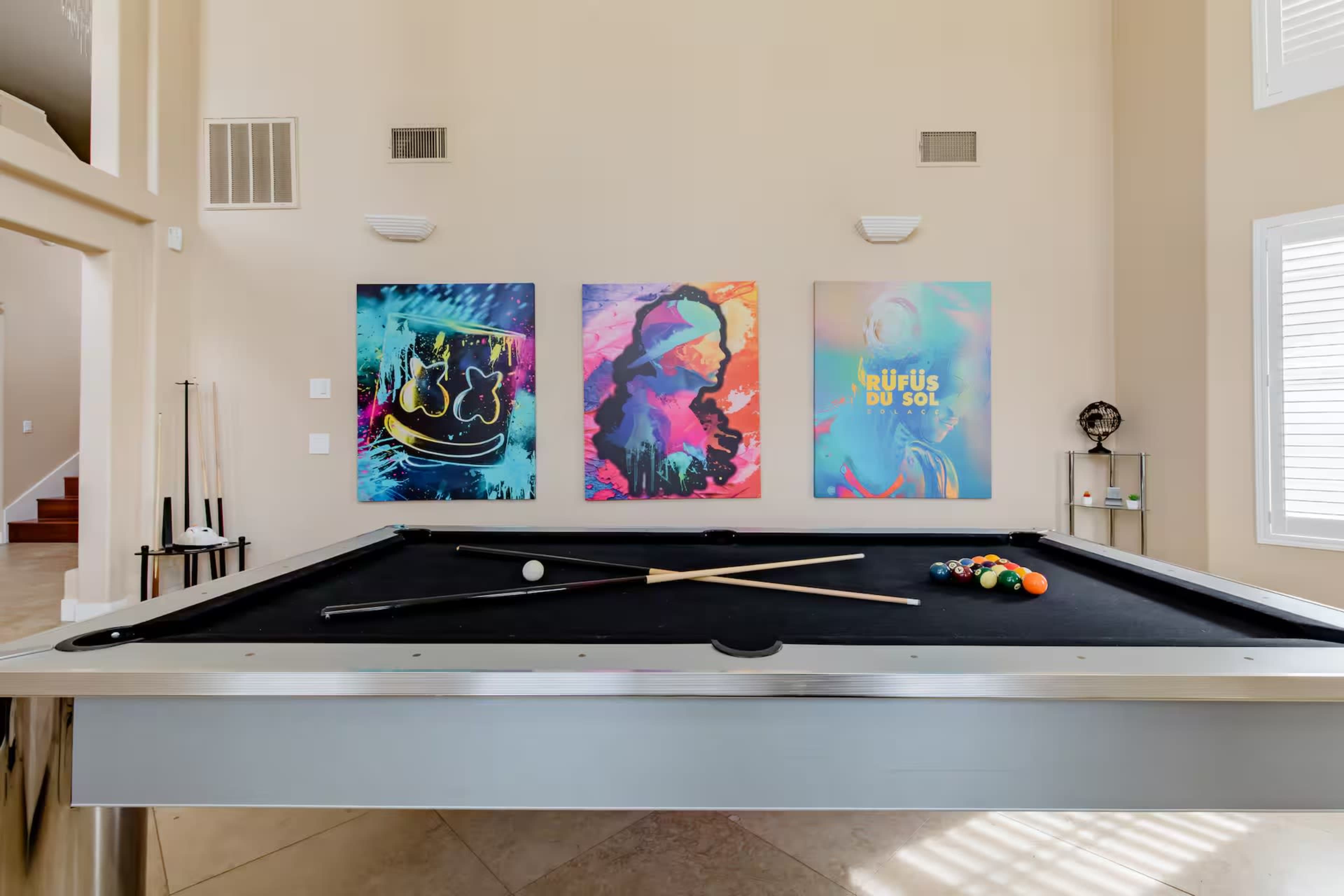 A billiard table with cues and balls is positioned in a room featuring three colorful abstract paintings on the wall.