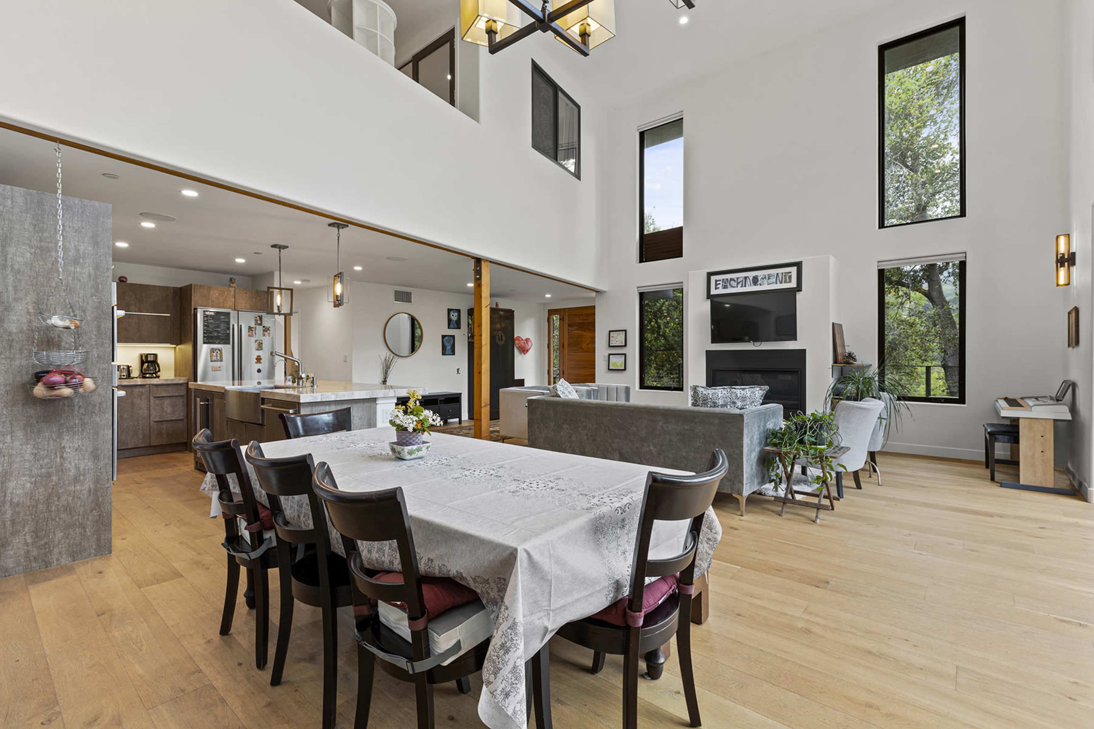 The image shows a modern, open-concept living and dining area with large windows, a dining table set for six, and a living room featuring a sofa and a television.