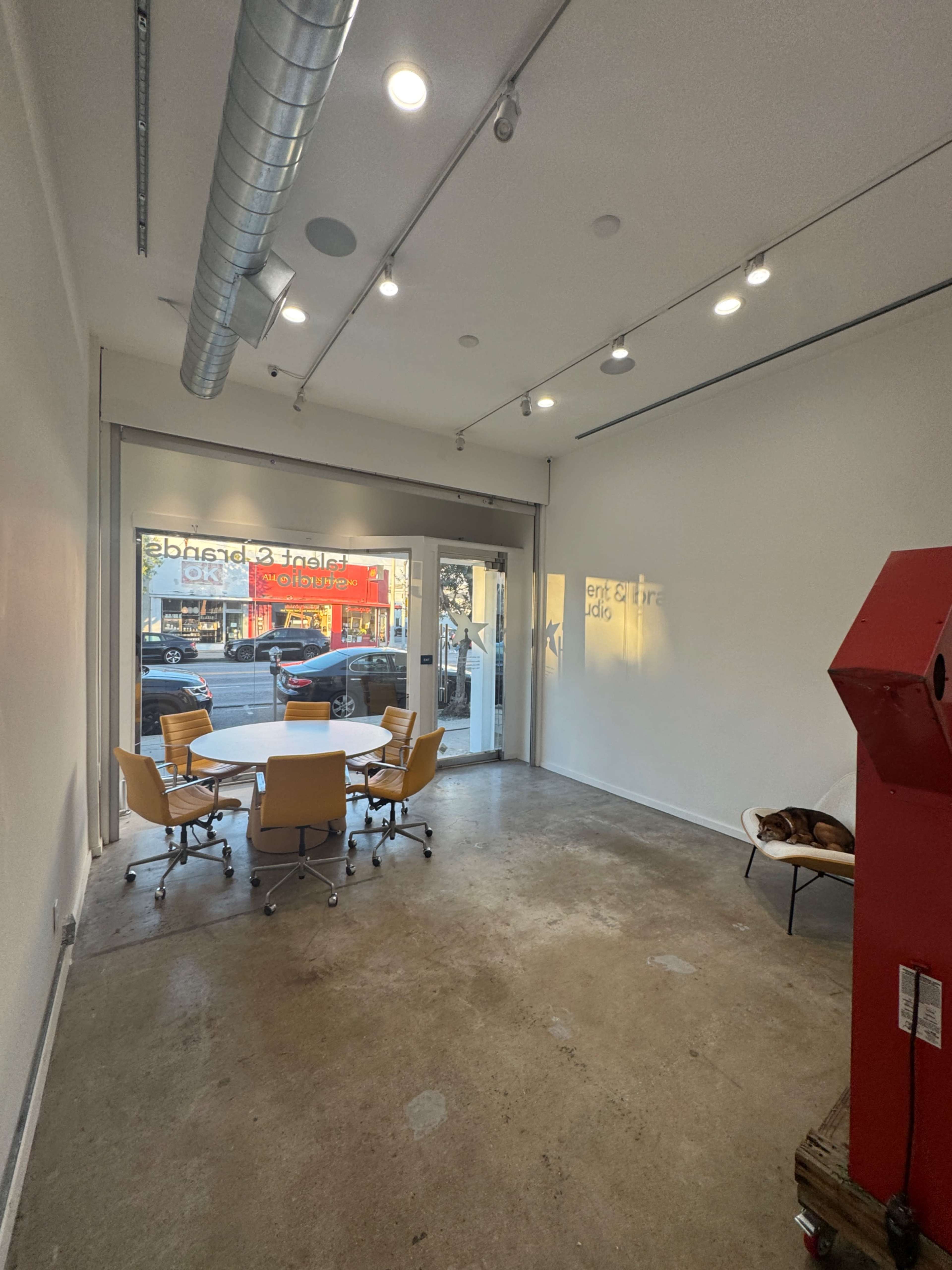 The image depicts a modern, empty office space with a round table and six chairs, large glass doors, and a visible red machine in one corner.