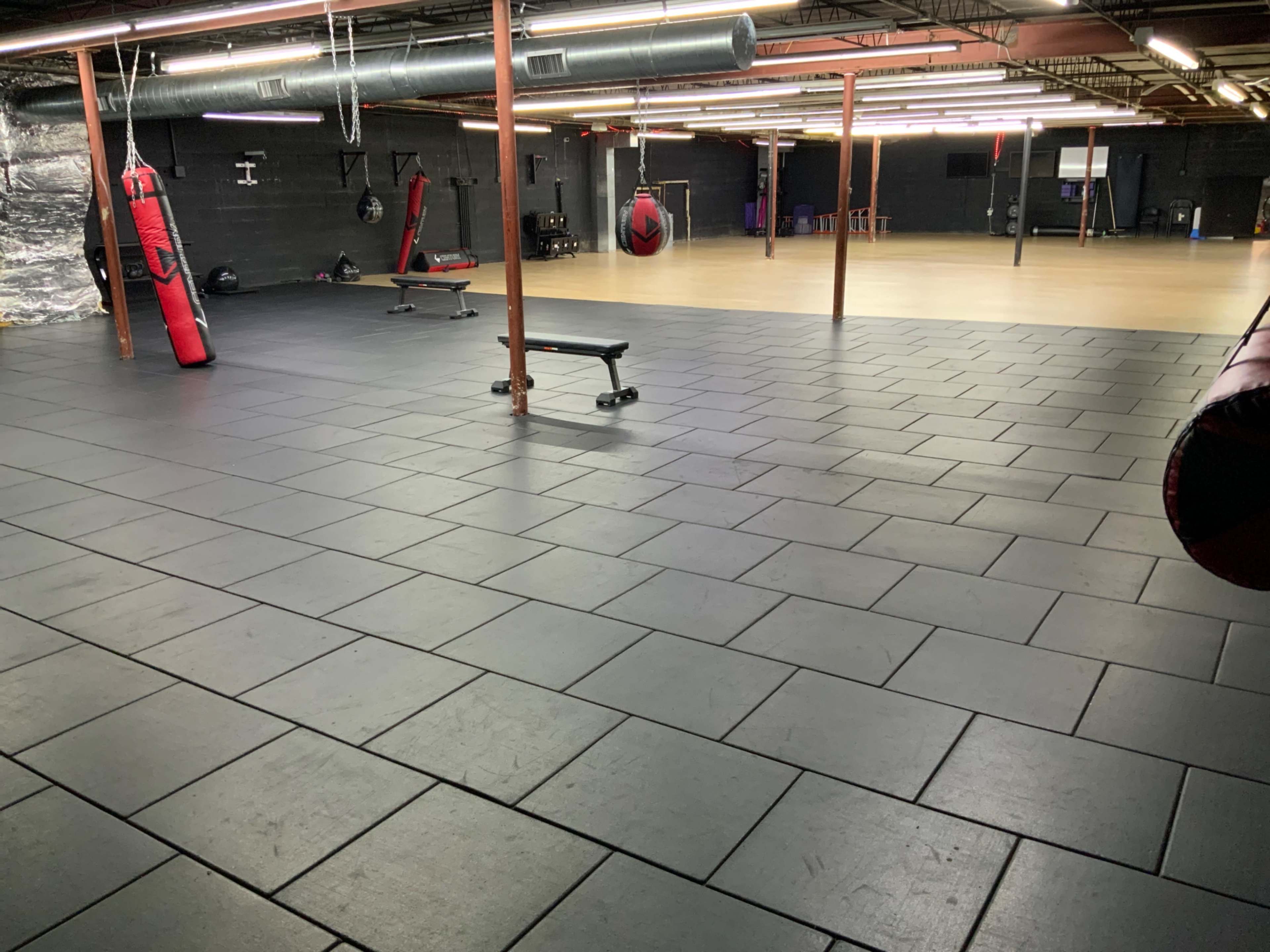 The image shows a spacious gym floor covered with black mats, featuring various fitness equipment and punching bags in a dimly lit area.