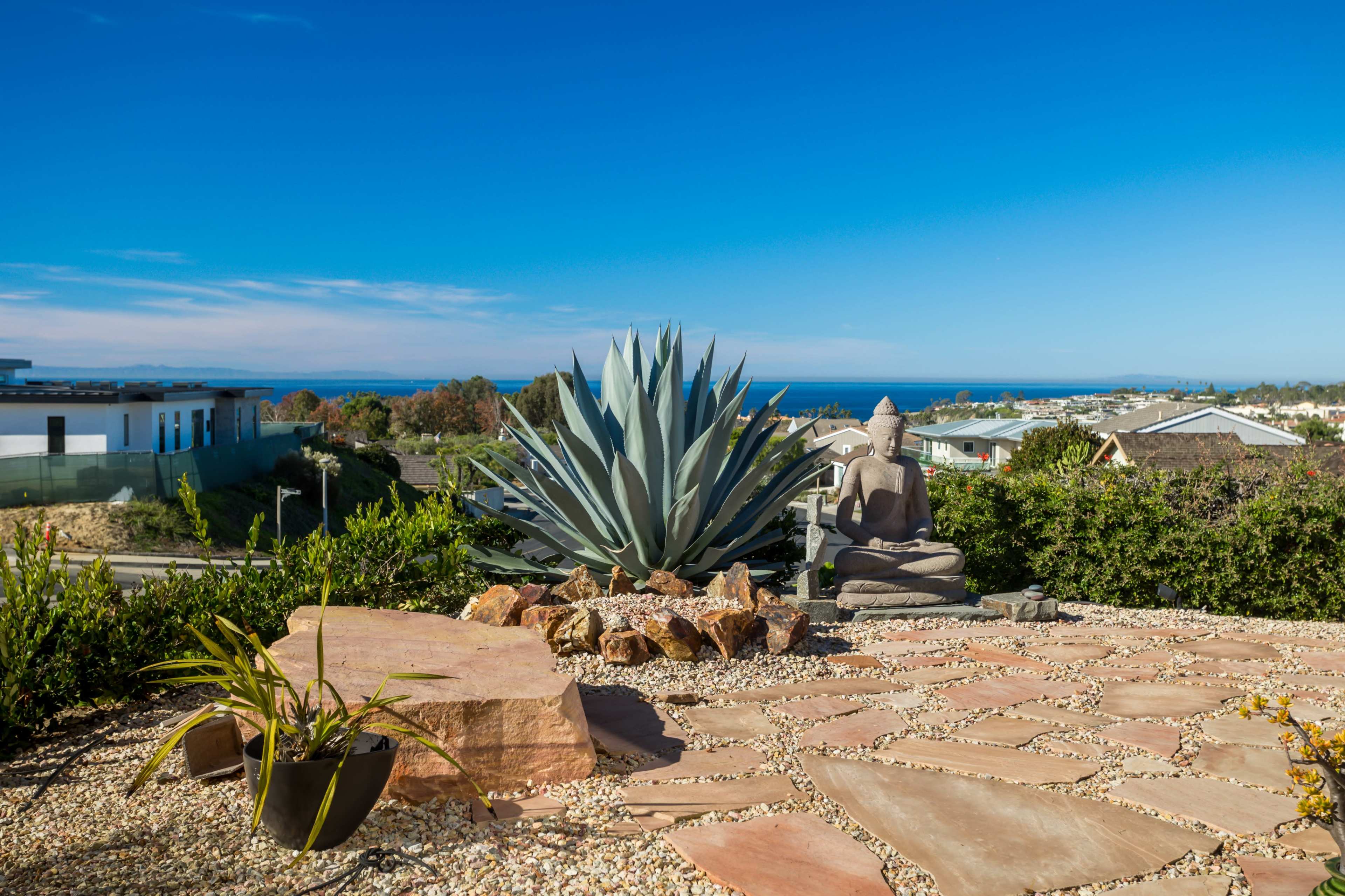 The scene features a landscaped yard with an agave plant and a Buddha statue overlooking the ocean and nearby houses under a clear blue sky.