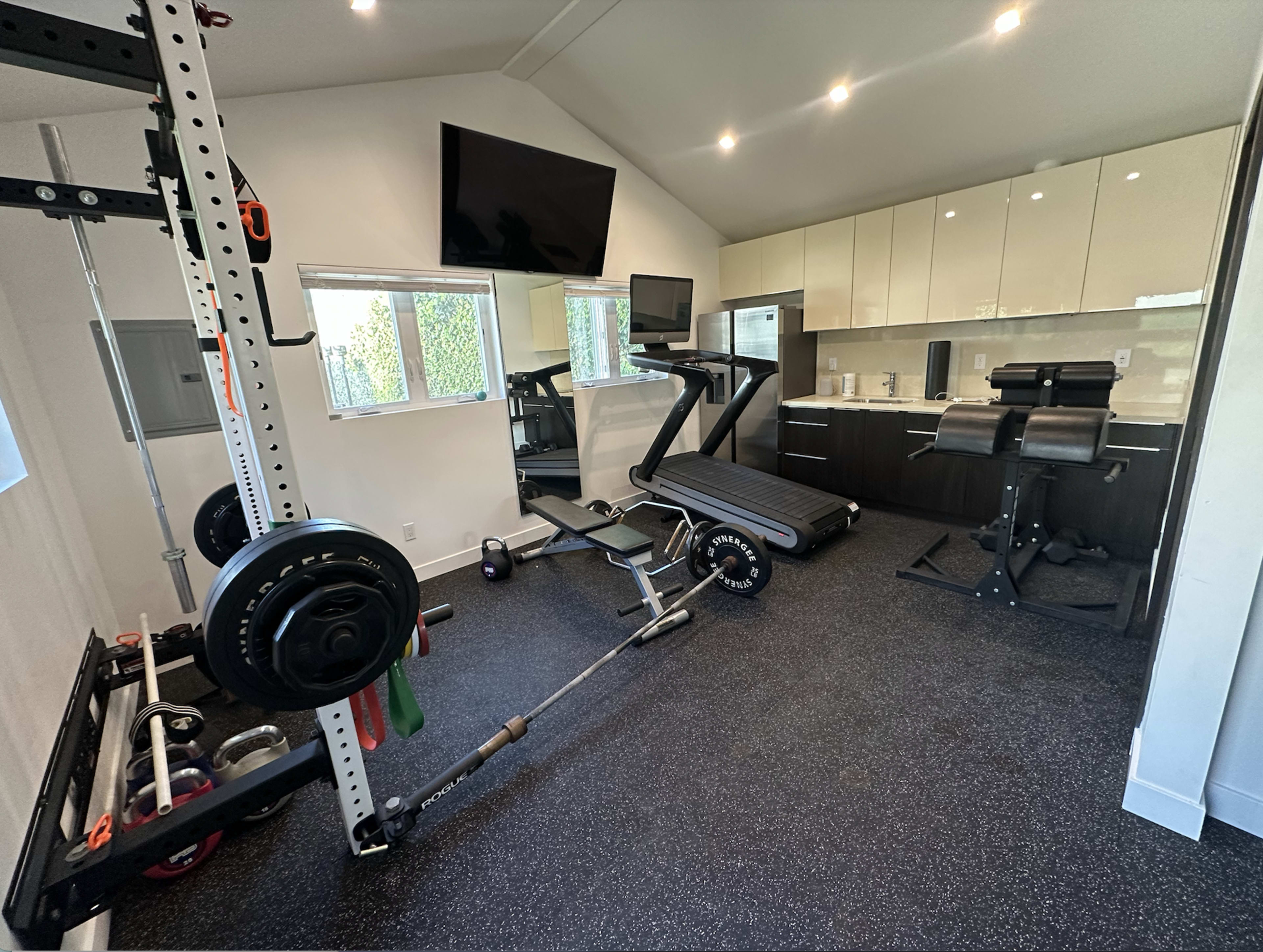 The image shows a home gym featuring a treadmill, weightlifting equipment, a multi-purpose bench, and a television mounted on the wall.