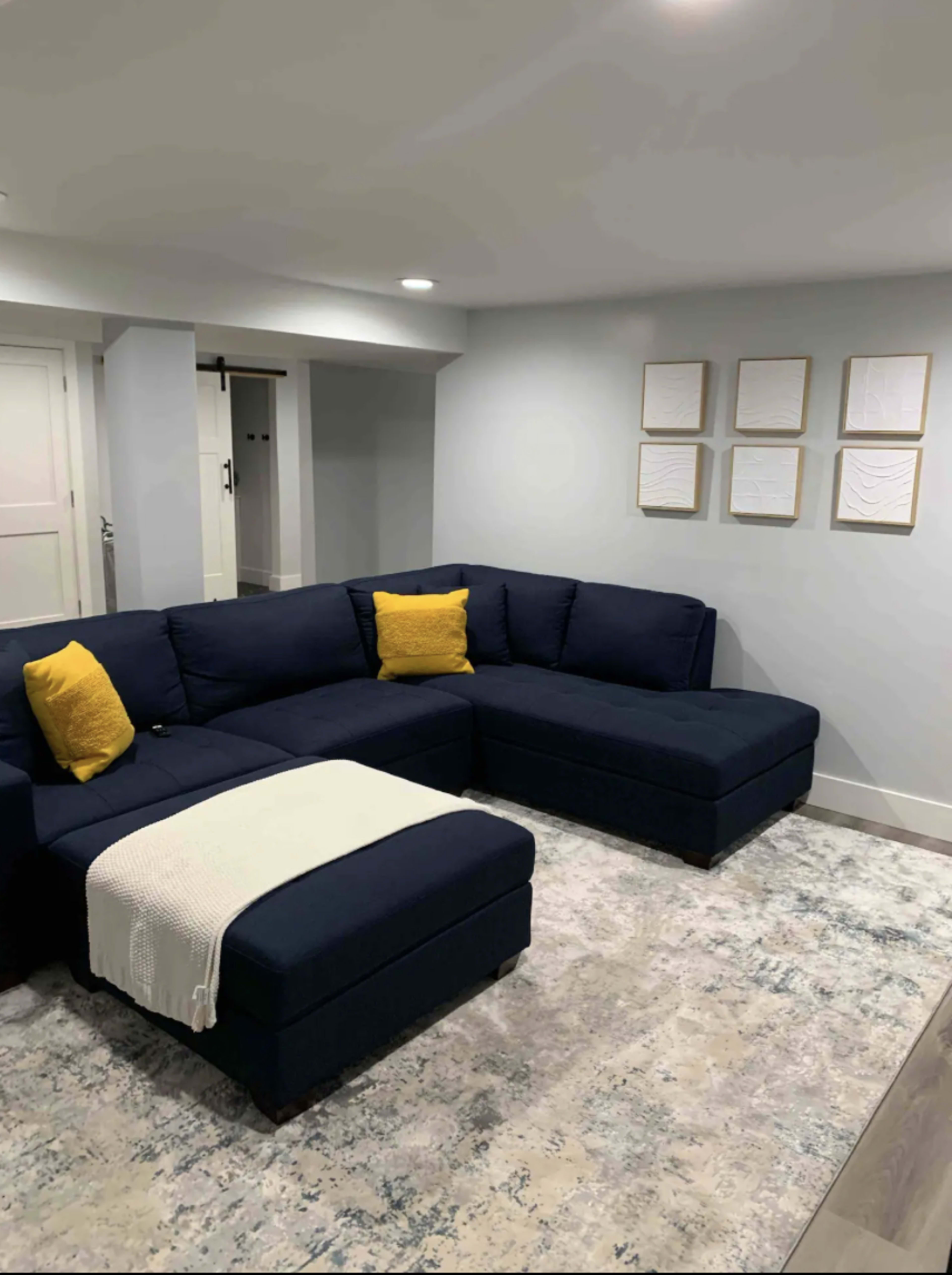 A modern basement living area features a dark blue sectional sofa with yellow accent pillows, a light throw blanket, and framed artworks on the wall.