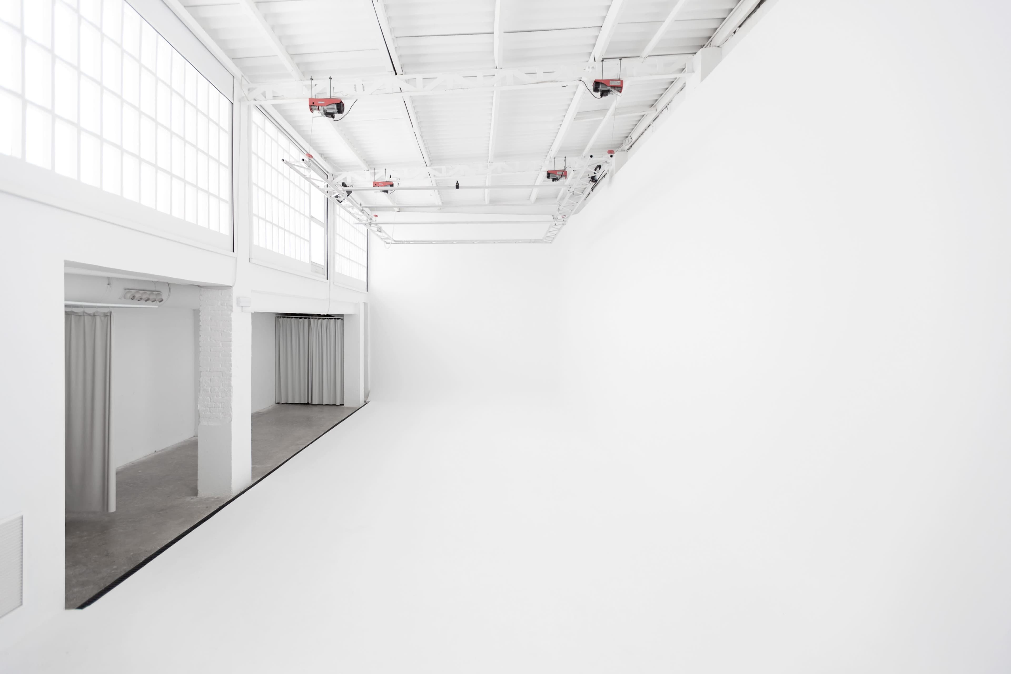 The image shows a spacious, minimally decorated white room with large windows and a polished concrete floor.