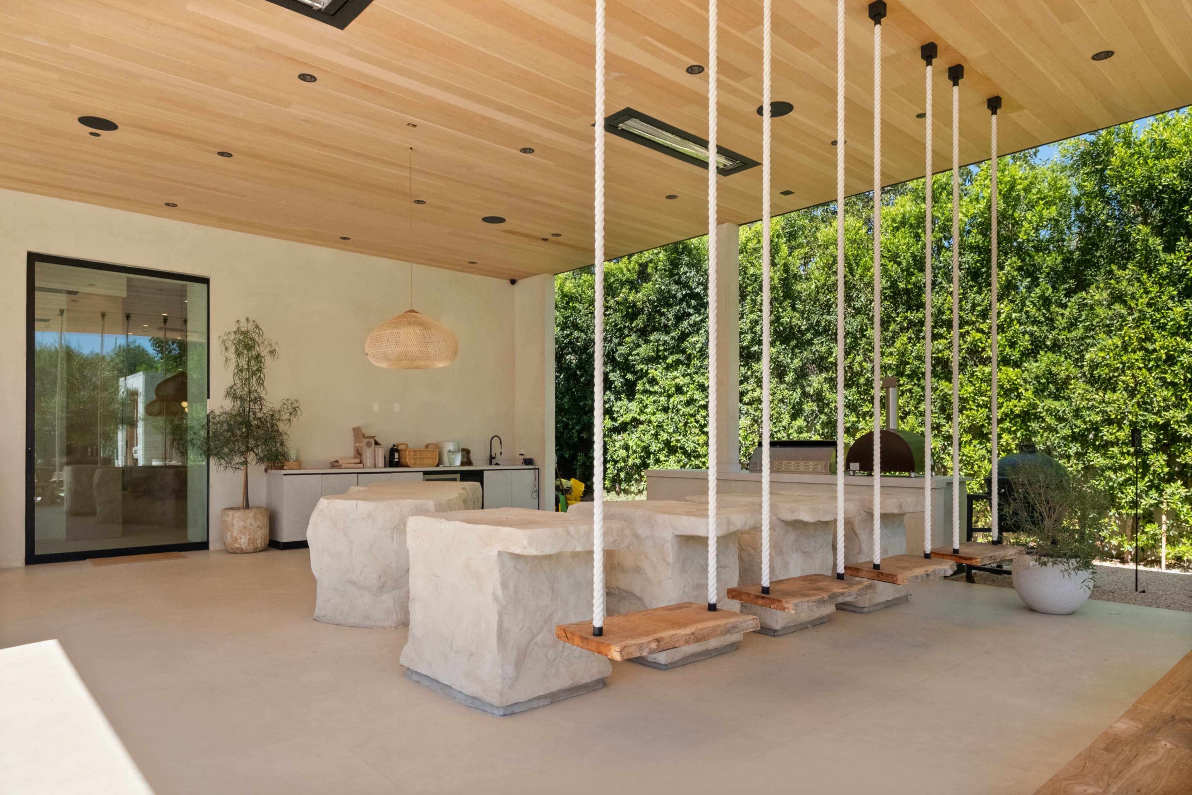 The image shows a modern outdoor space featuring stone seating, wooden swings, and a kitchenette with large glass doors opening to greenery.