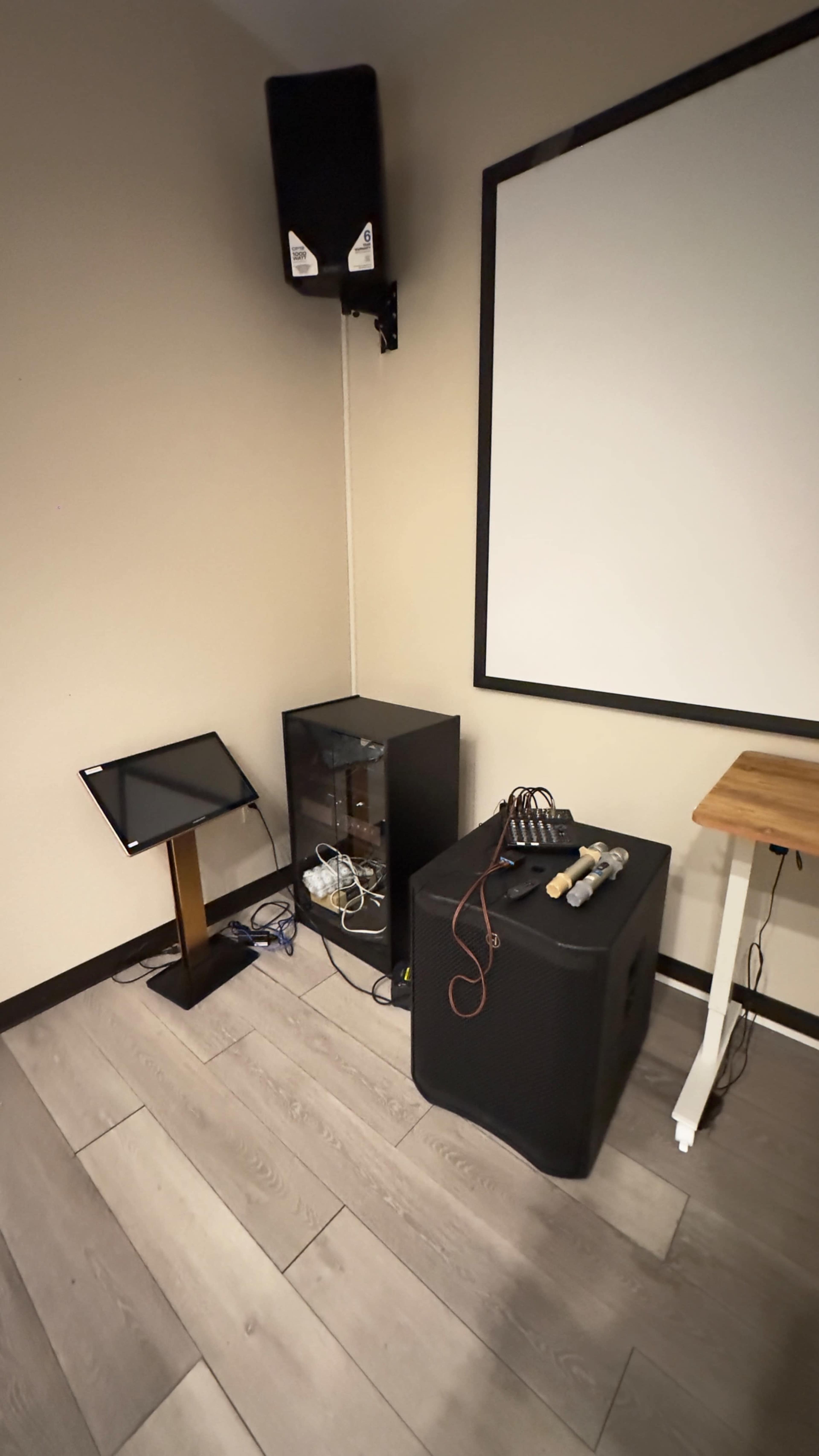 The image shows a corner of a room with a speaker mounted on the wall, a black cabinet containing electronic equipment, and a subwoofer on the floor next to a small table.