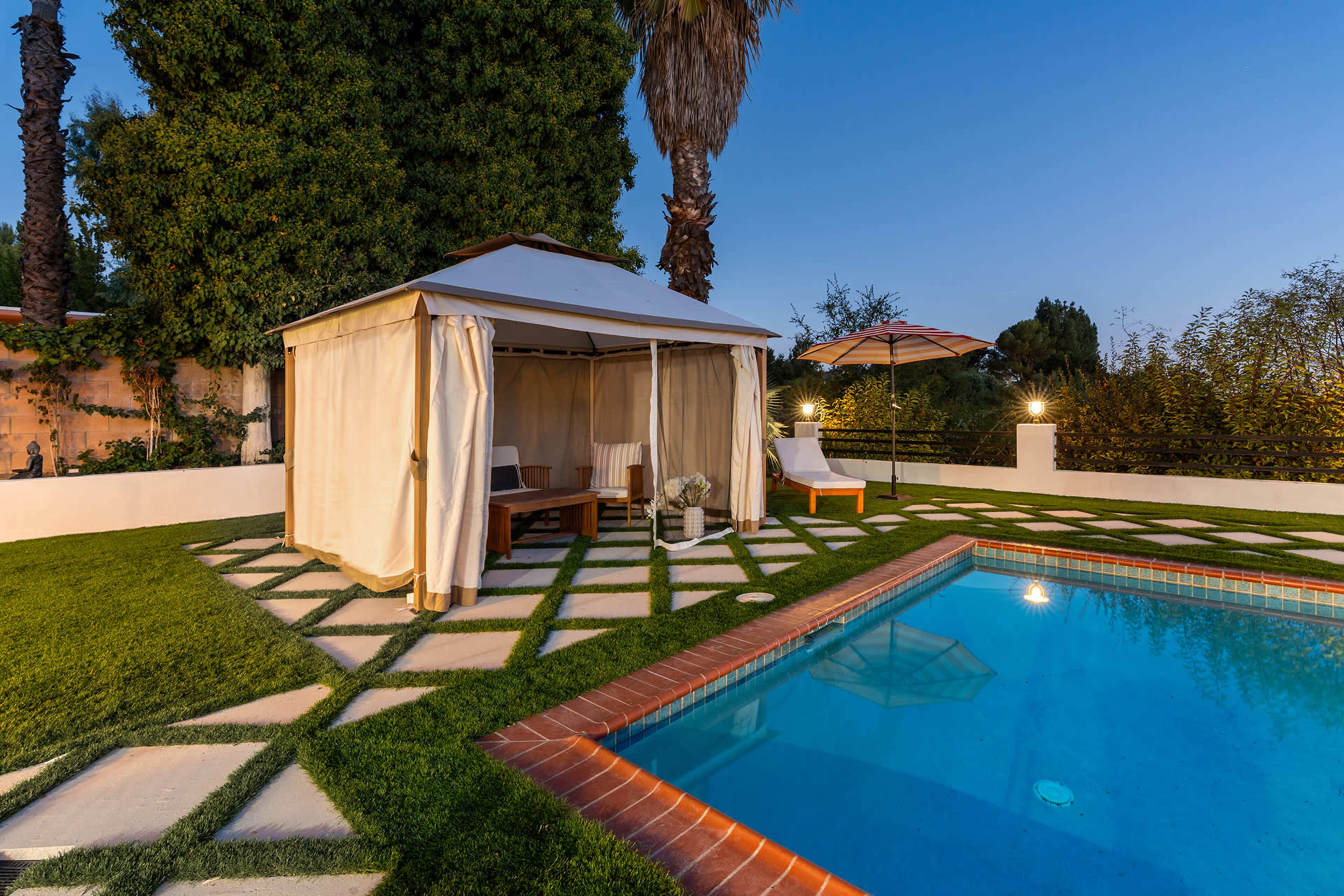 The image shows a backyard with a swimming pool, a gazebo with curtains, and lounge chairs, all surrounded by well-maintained grass and landscaping.