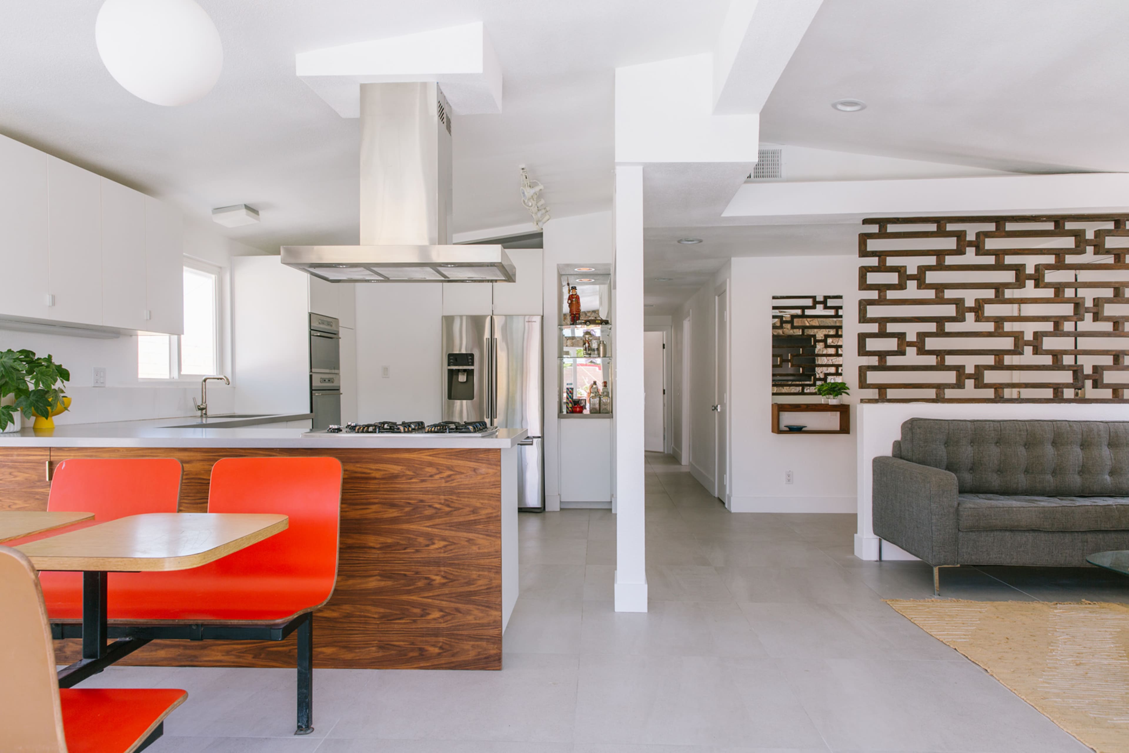 A modern kitchen and living area showcase a stainless steel range hood, wooden cabinetry, and red dining chairs adjacent to a textured wall panel.