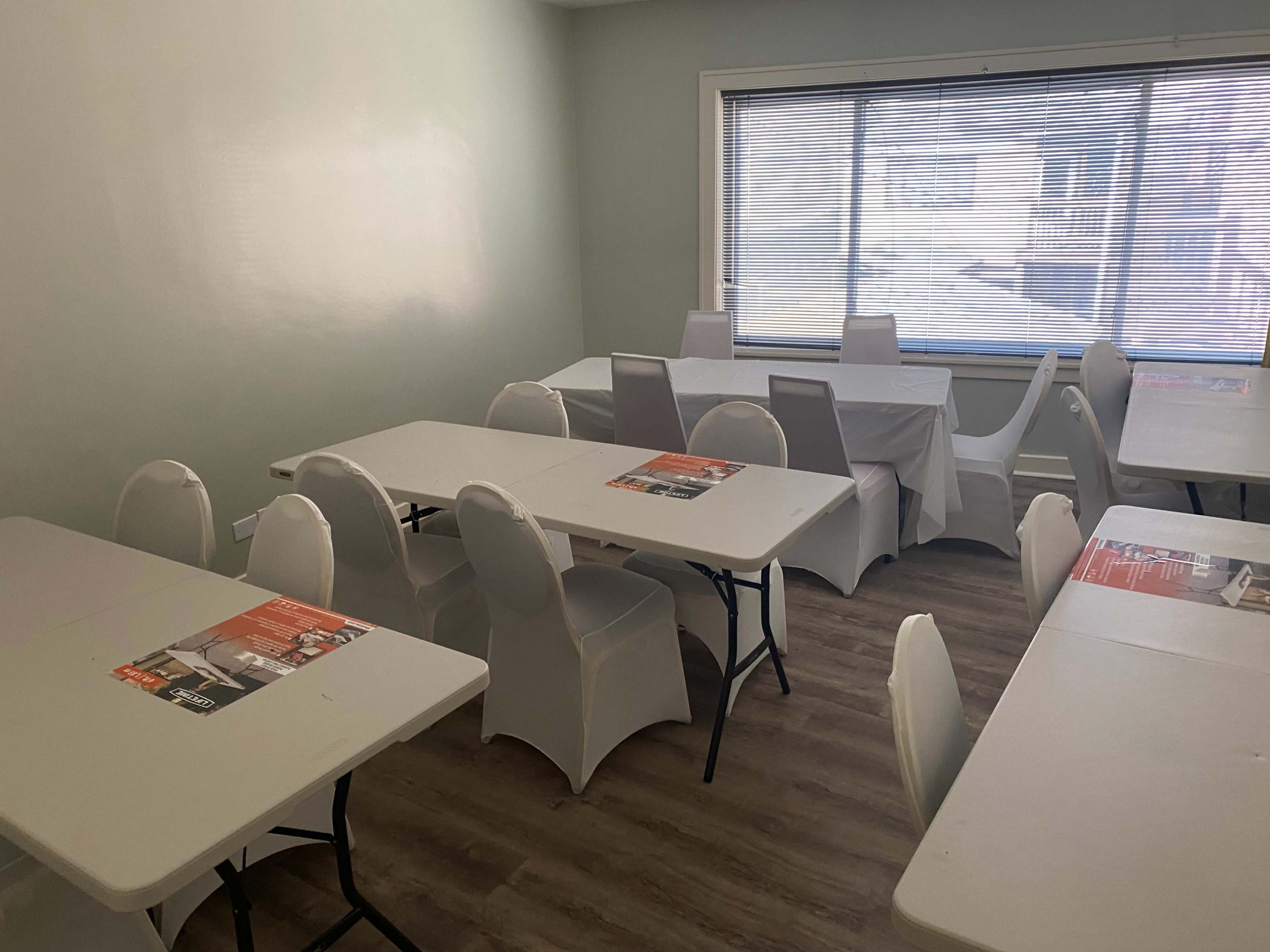 A vacant meeting room with several tables and chairs arranged, and large windows letting in natural light.