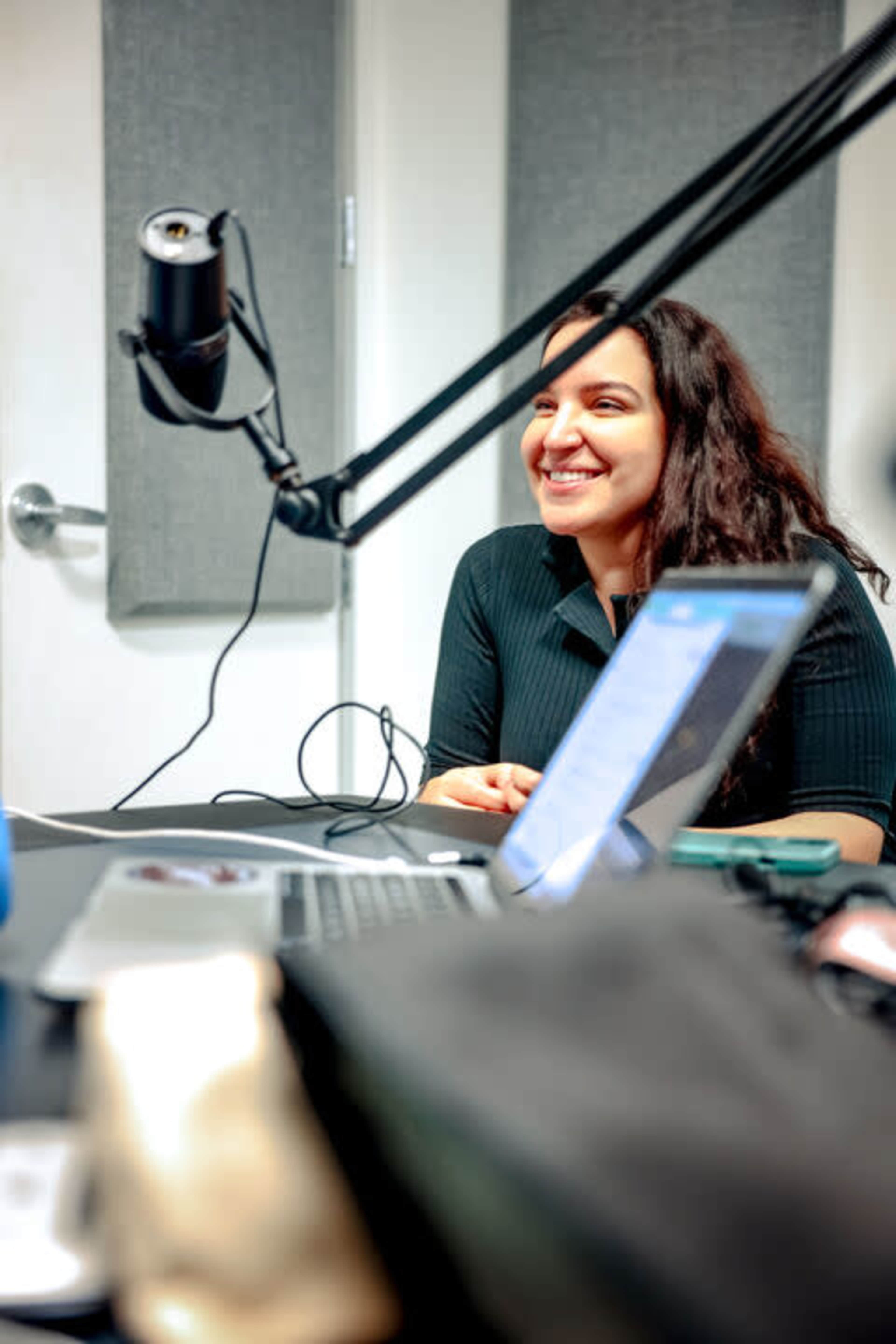 A woman with long, dark hair is smiling while sitting at a table with a microphone and laptops in a recording studio.