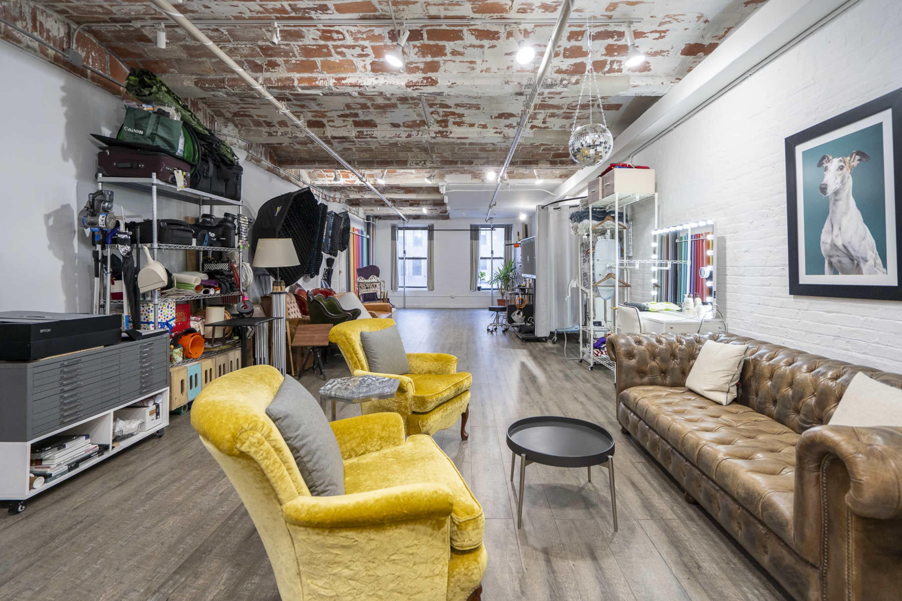 An open, well-lit interior space featuring a mix of vintage and modern furnishings, with a yellow armchair, a brown leather sofa, and various storage items along the walls.
