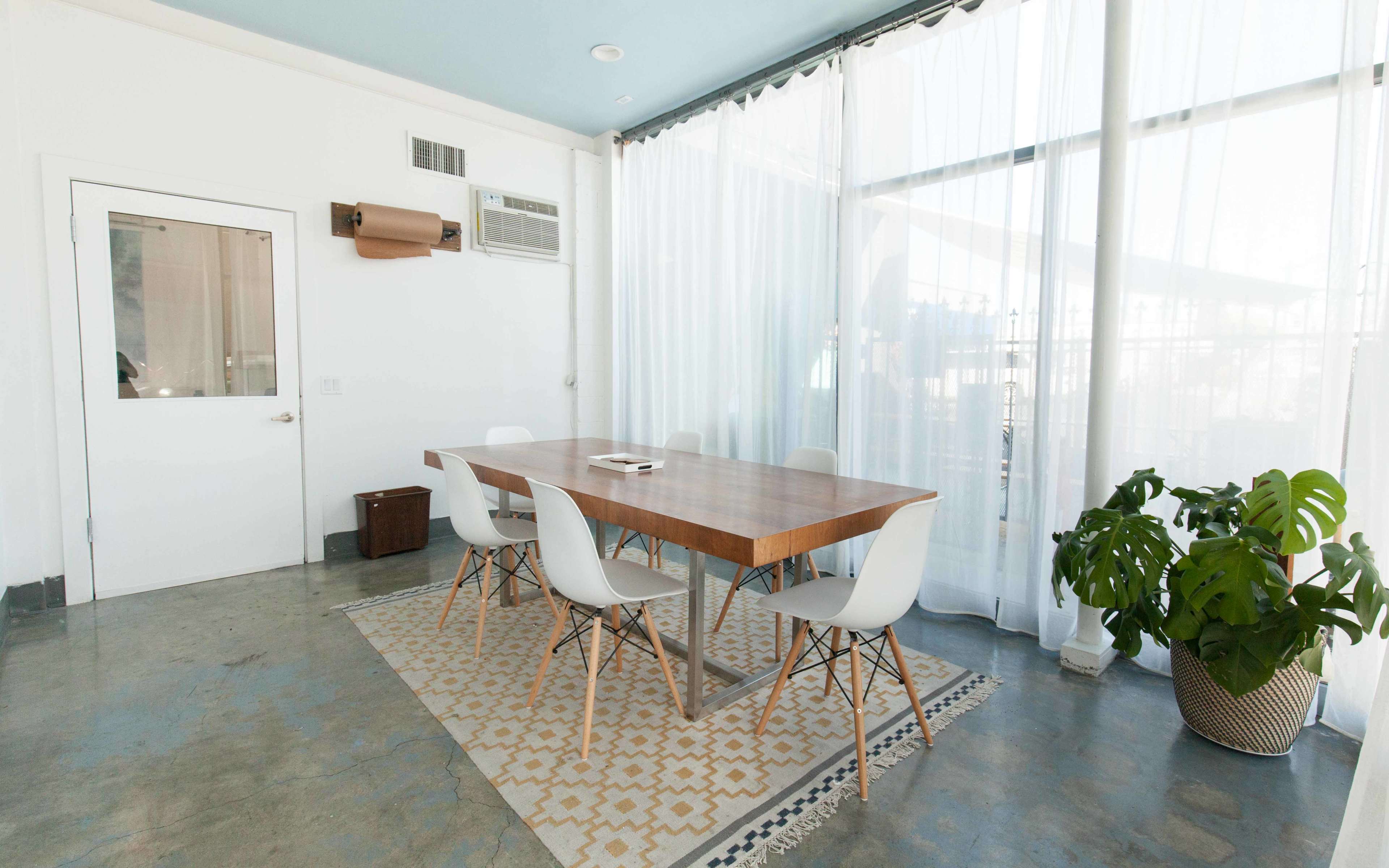 A bright and airy meeting room features a wooden table surrounded by four white chairs, with large windows draped in sheer curtains and a potted plant in the corner.