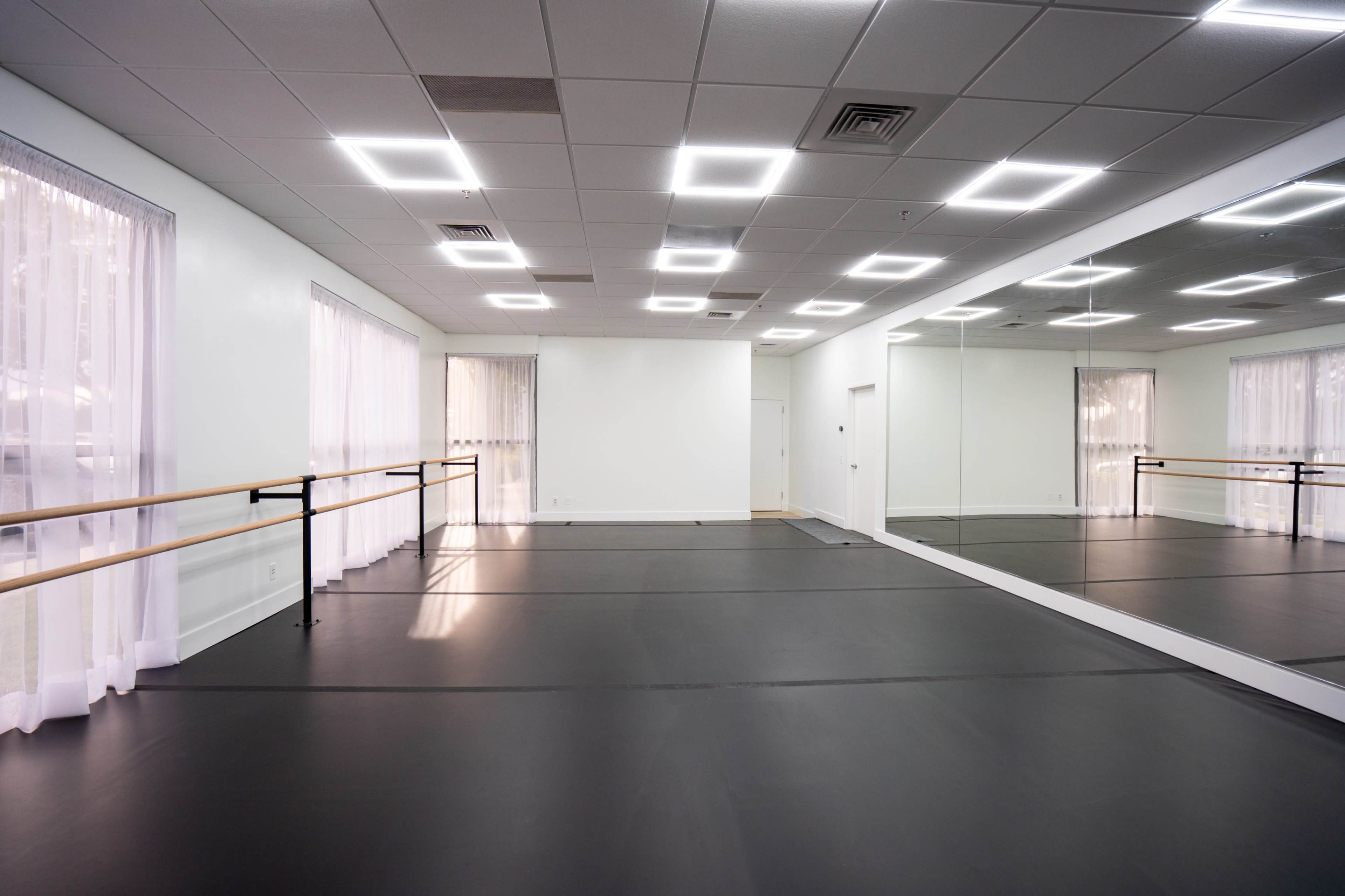 The image shows a spacious dance studio with a mirrored wall, wooden ballet bar, and large windows covered with sheer curtains.