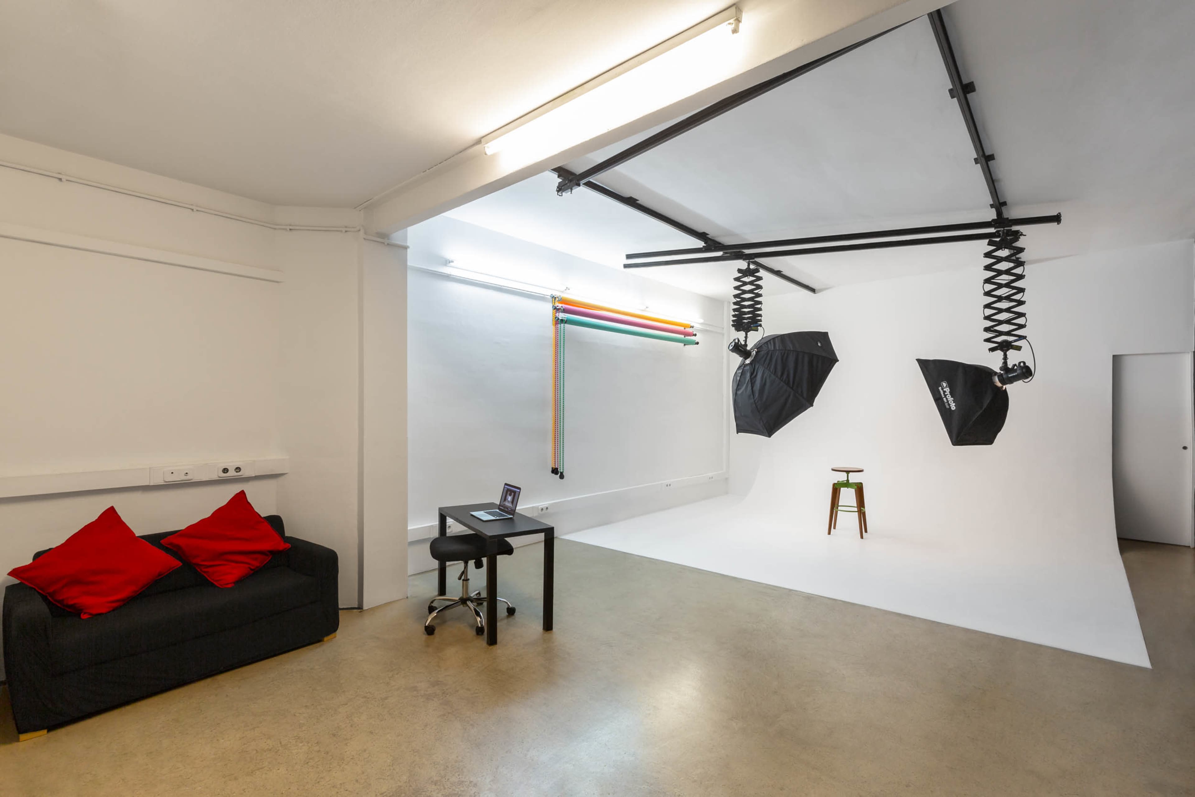 The image shows a photography studio featuring a white backdrop, two large softbox lights, a stool, and a black sofa with red cushions.