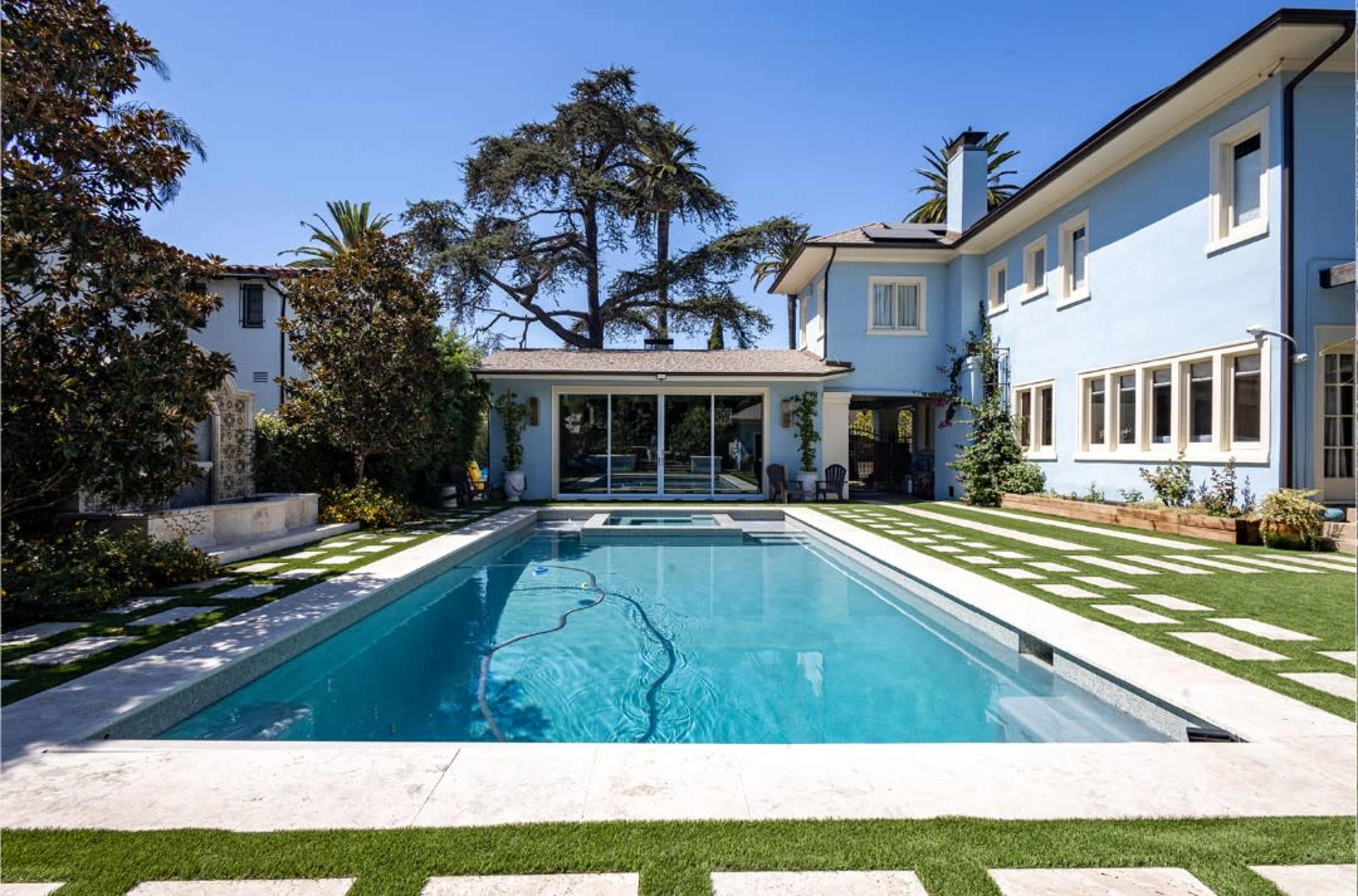 The image depicts a modern blue house with a rectangular swimming pool, surrounded by a manicured lawn and decorative landscaping.