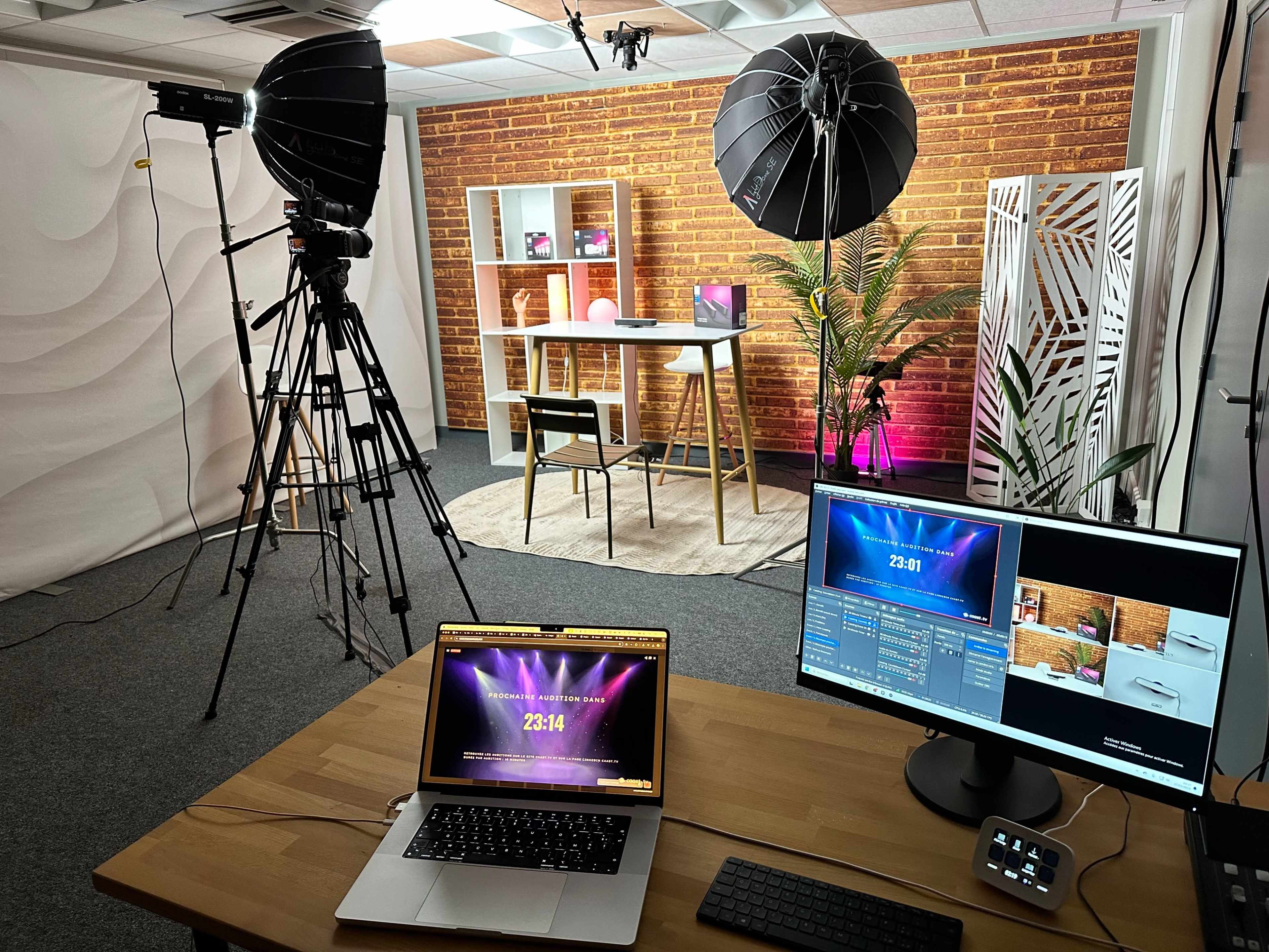 The image shows a professional video setup with two large softbox lights, a tripod, a desk with a plant, and two computer monitors displaying a countdown timer, all set against a brick wall backdrop.