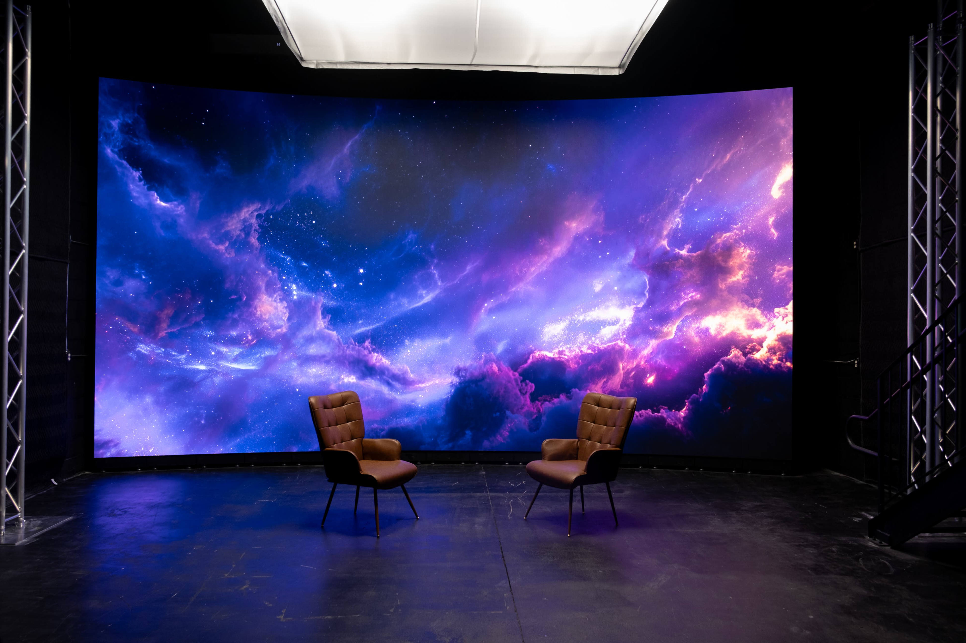 The scene features two brown chairs positioned in front of a large, curved screen displaying a colorful cosmic backdrop of purple and blue nebulae.