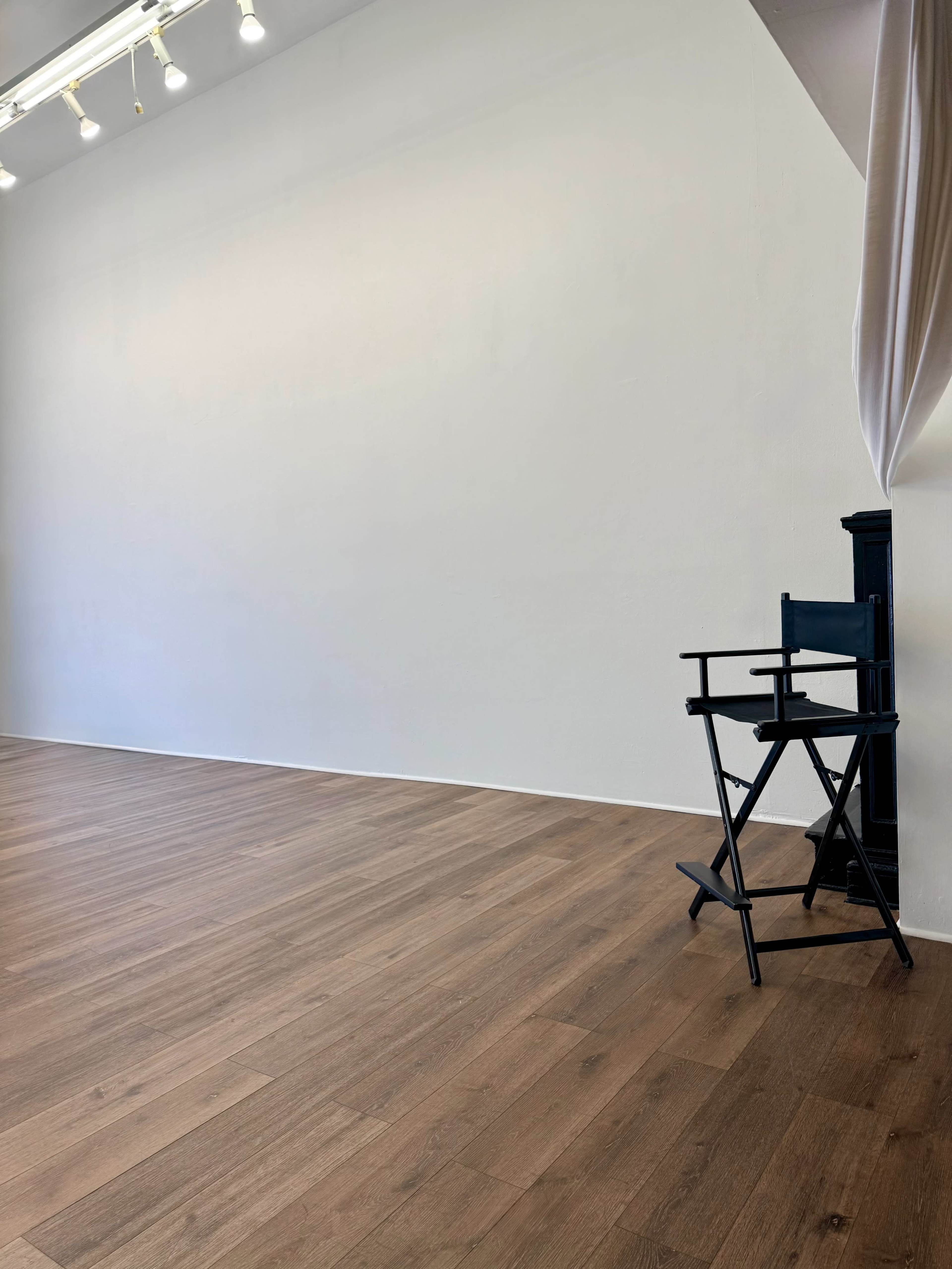 The image shows an empty room with wooden flooring, a plain white wall, and a black director's chair positioned in the corner.