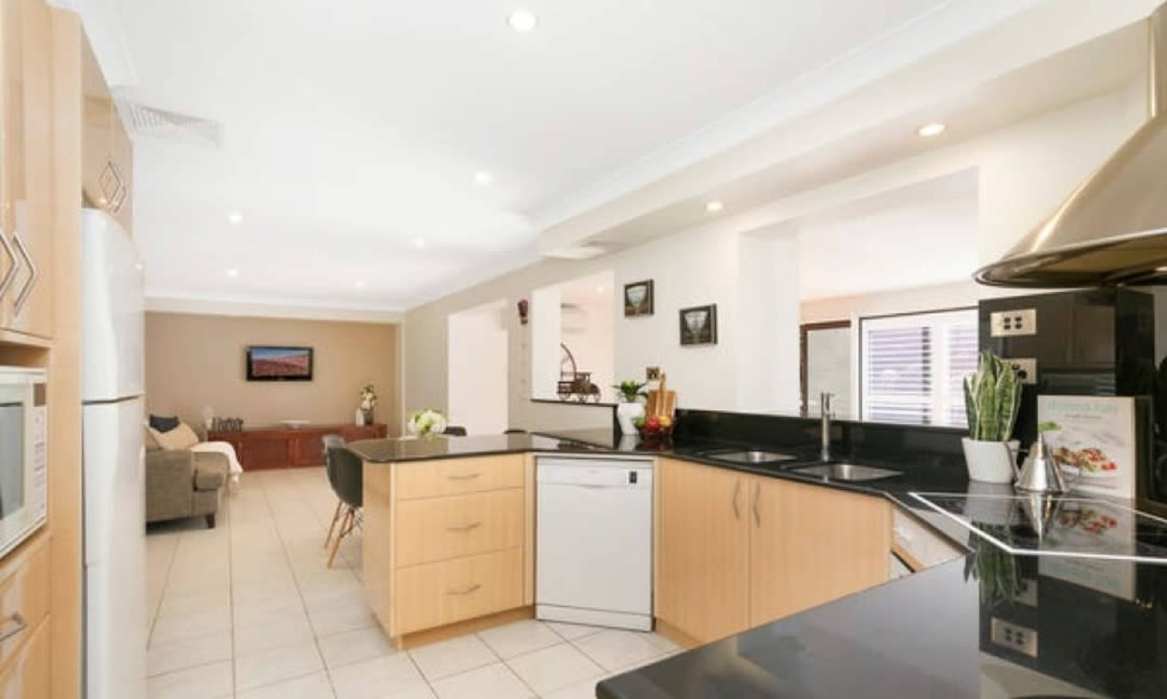 A modern kitchen features light wood cabinetry, black countertops, and stainless steel appliances, with a view into a living area in the background.
