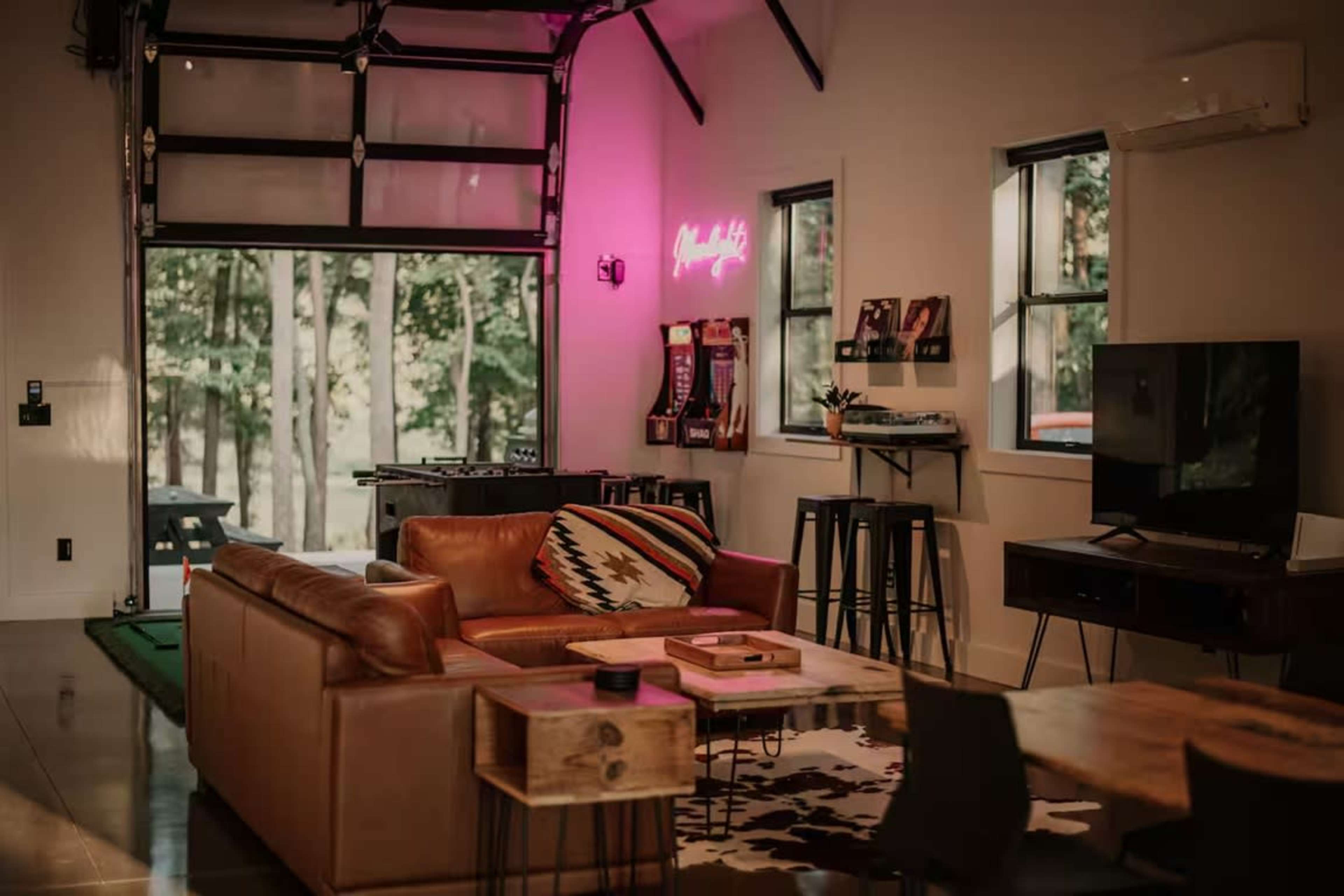The image shows a modern living space with a large brown leather sofa, a wooden coffee table, and a flat-screen TV, featuring large windows that overlook a wooded area.