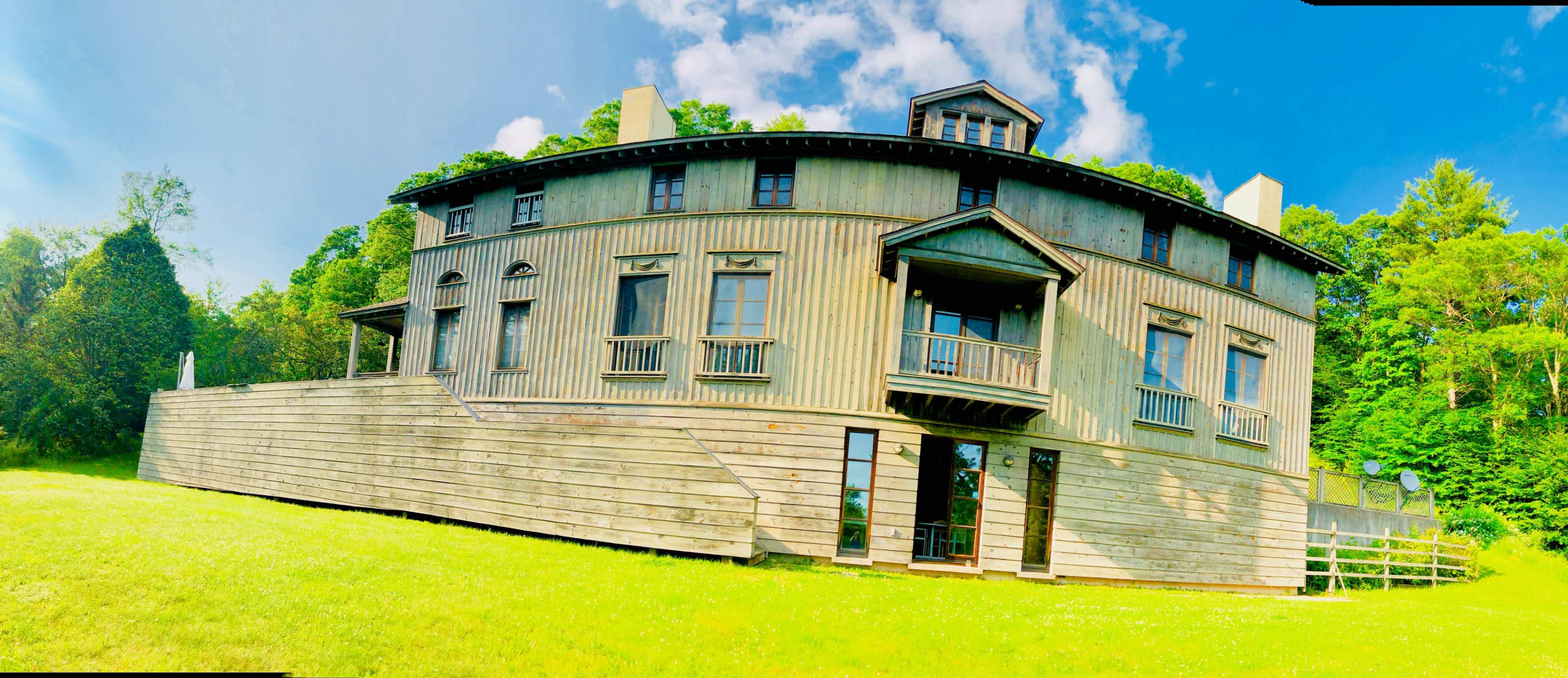 A two-story, wooden house with a unique round design, large windows, and a sloped lawn in front, surrounded by trees and a blue sky.