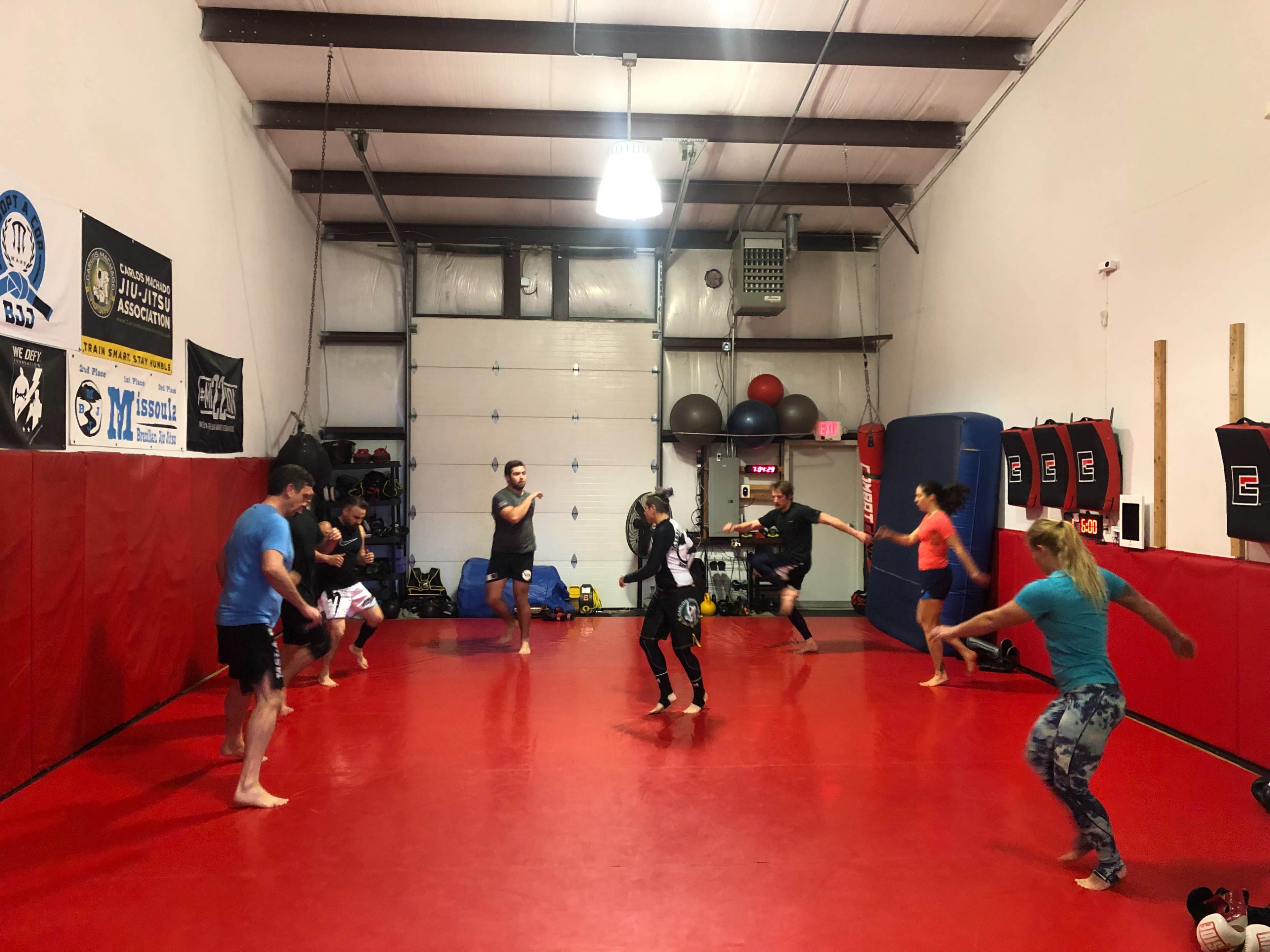 A group of people practice martial arts in a dojo with red mats and various training equipment.
