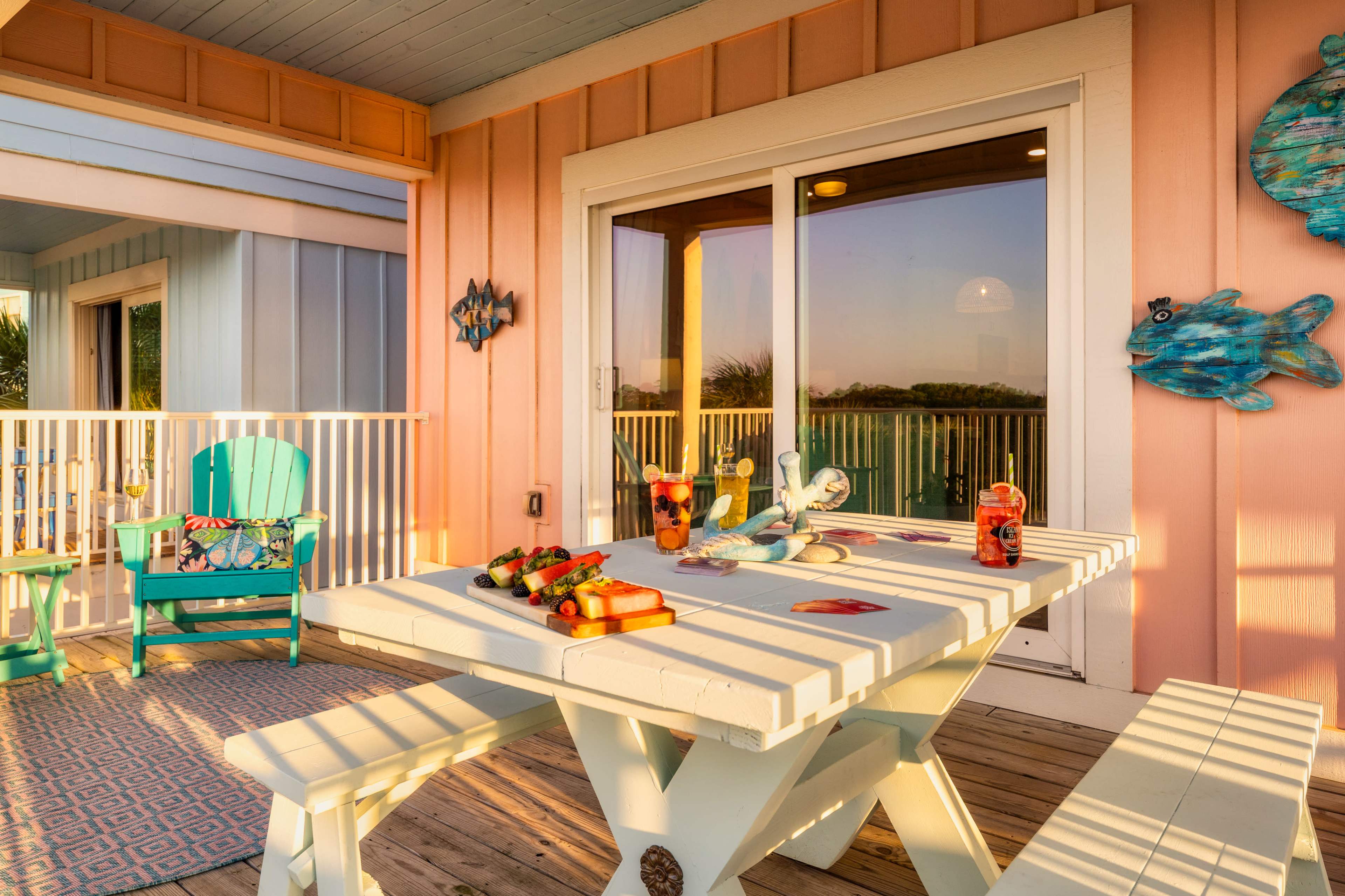 A brightly colored patio features a picnic table, decorated with snacks and drinks, surrounded by colorful chairs and coastal-themed decor.