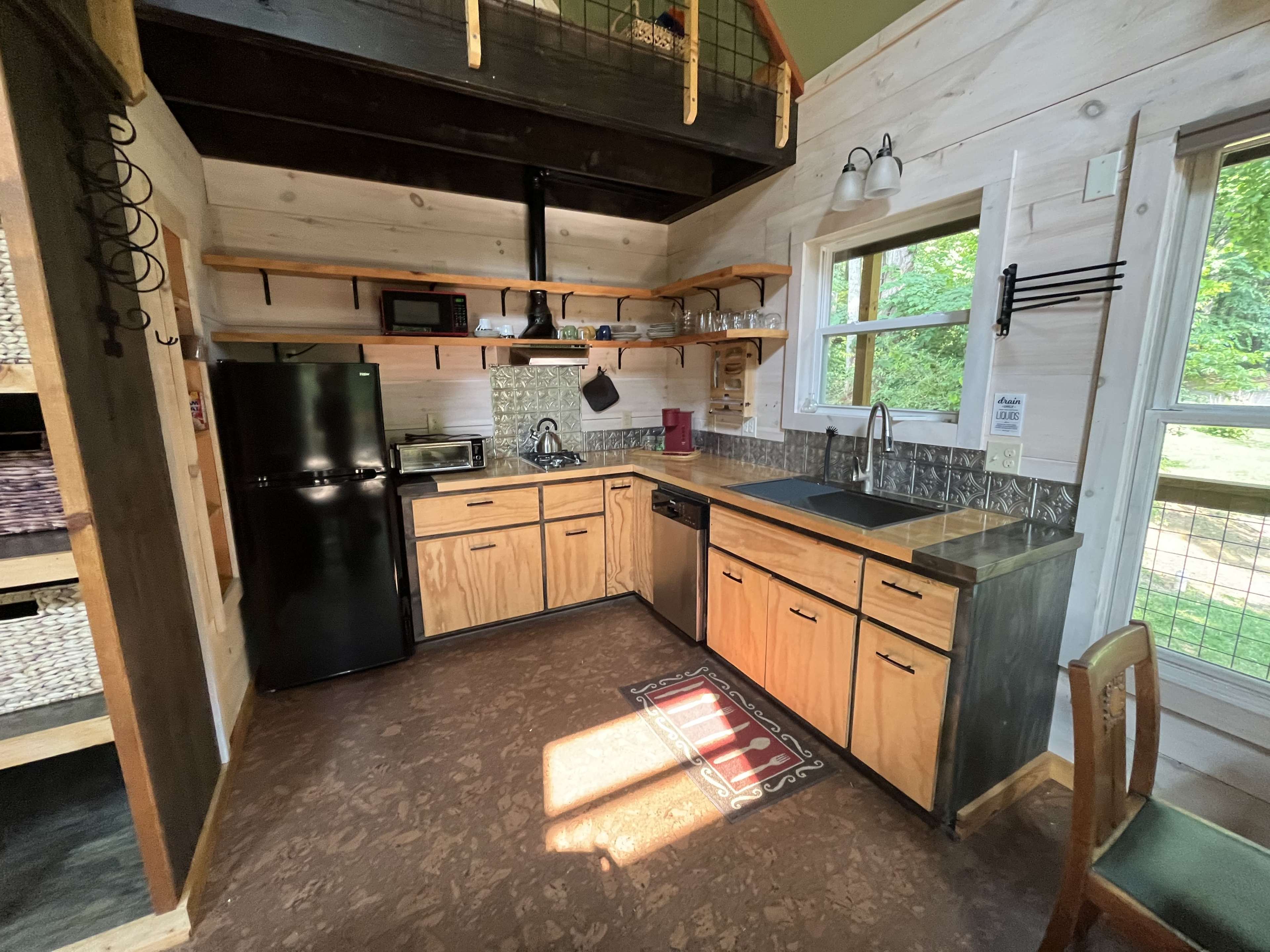The image shows a small kitchen in a wooden cabin featuring a black refrigerator, a microwave, a sink, cabinetry, and a window that provides natural light.