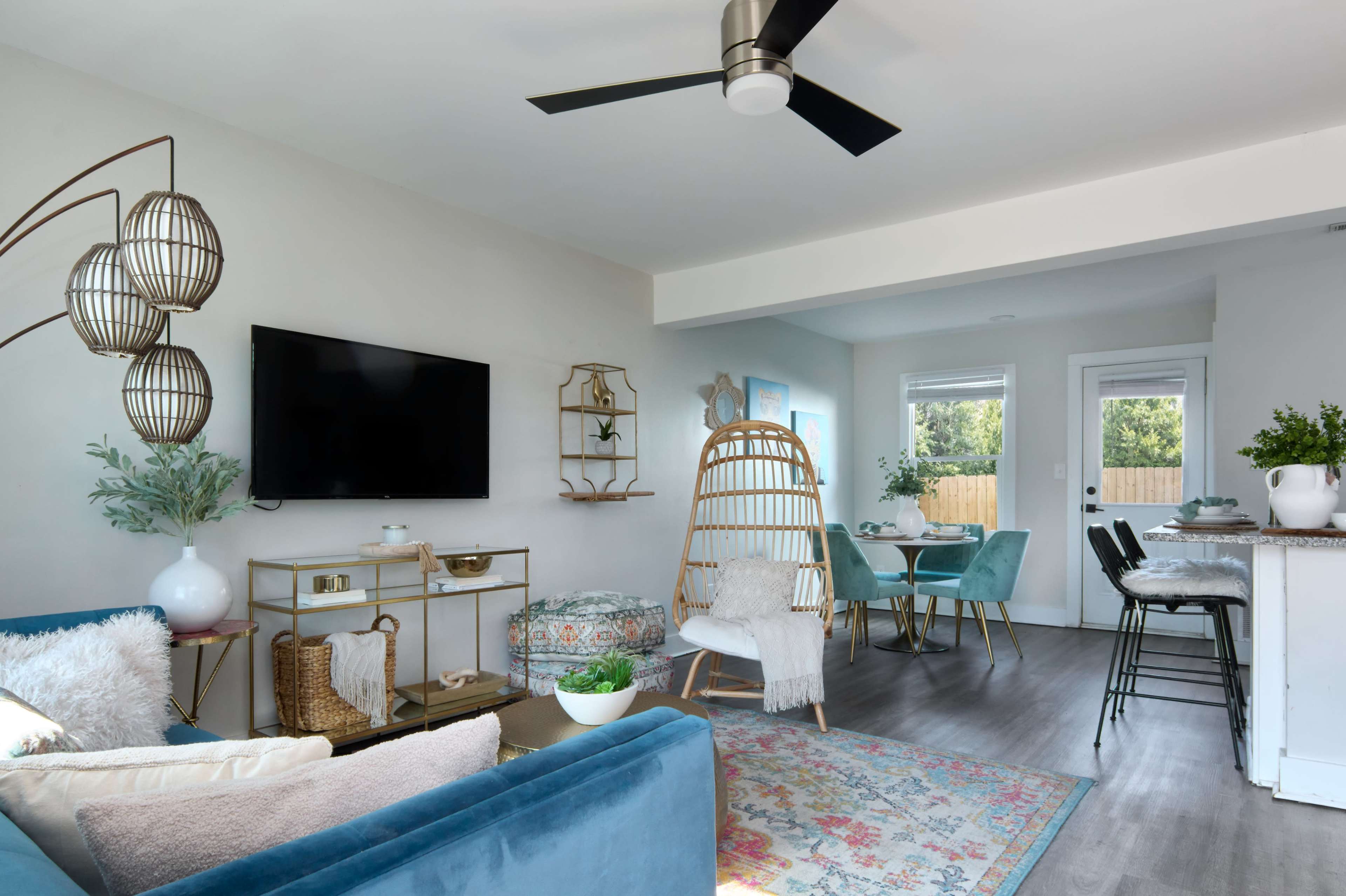 The image shows a modern living room featuring a teal couch, a wall-mounted television, a rattan hanging chair, and a dining area with black chairs and a table.