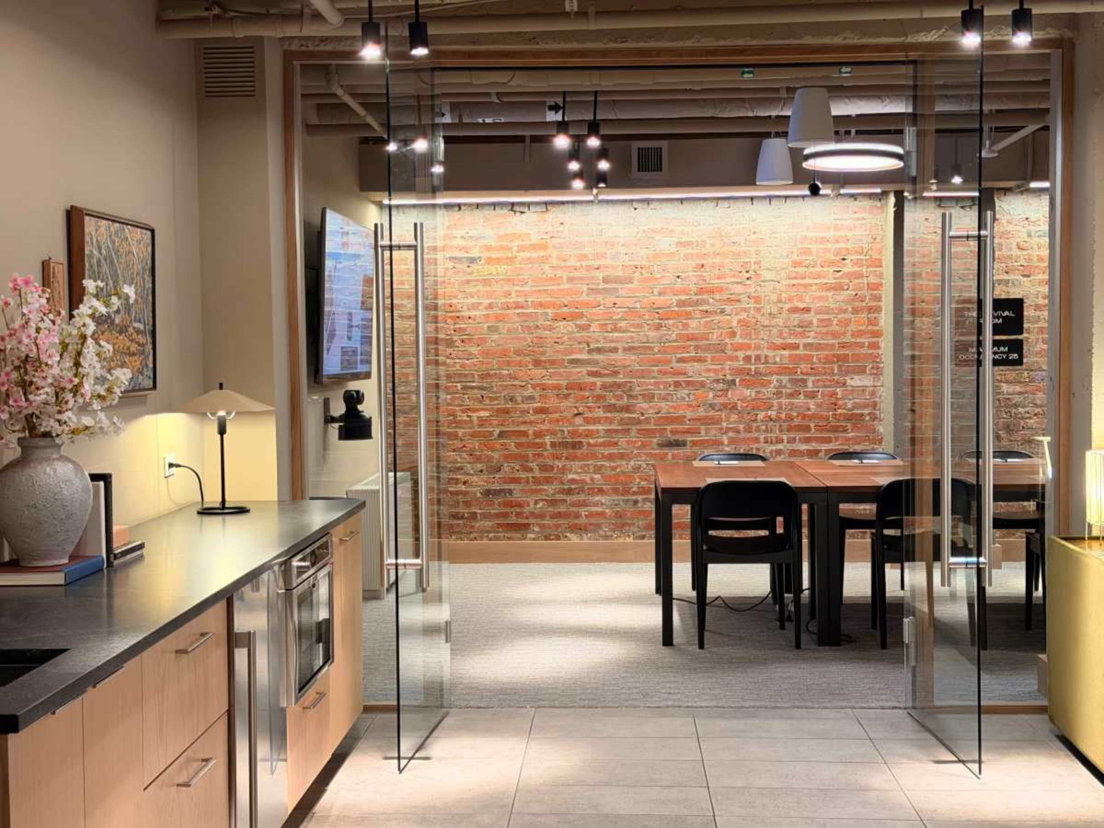 The image shows a modern office space with a kitchen area on the left, featuring a dark countertop and a decorative vase, and a meeting area with a wooden table and chairs against a brick wall.