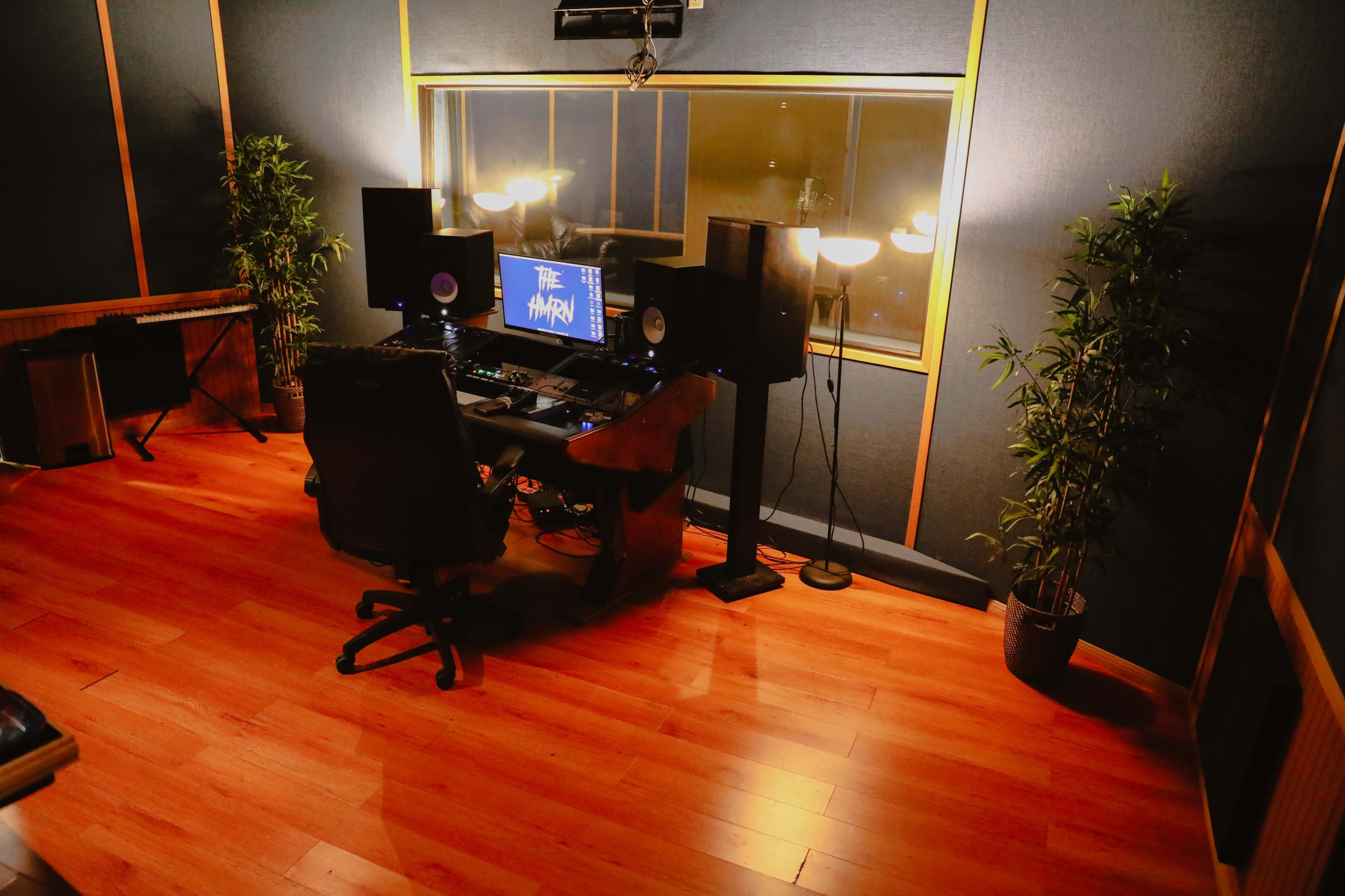The image shows a recording studio with a desk, two speakers, a computer monitor, and plants, all arranged in a well-lit space.