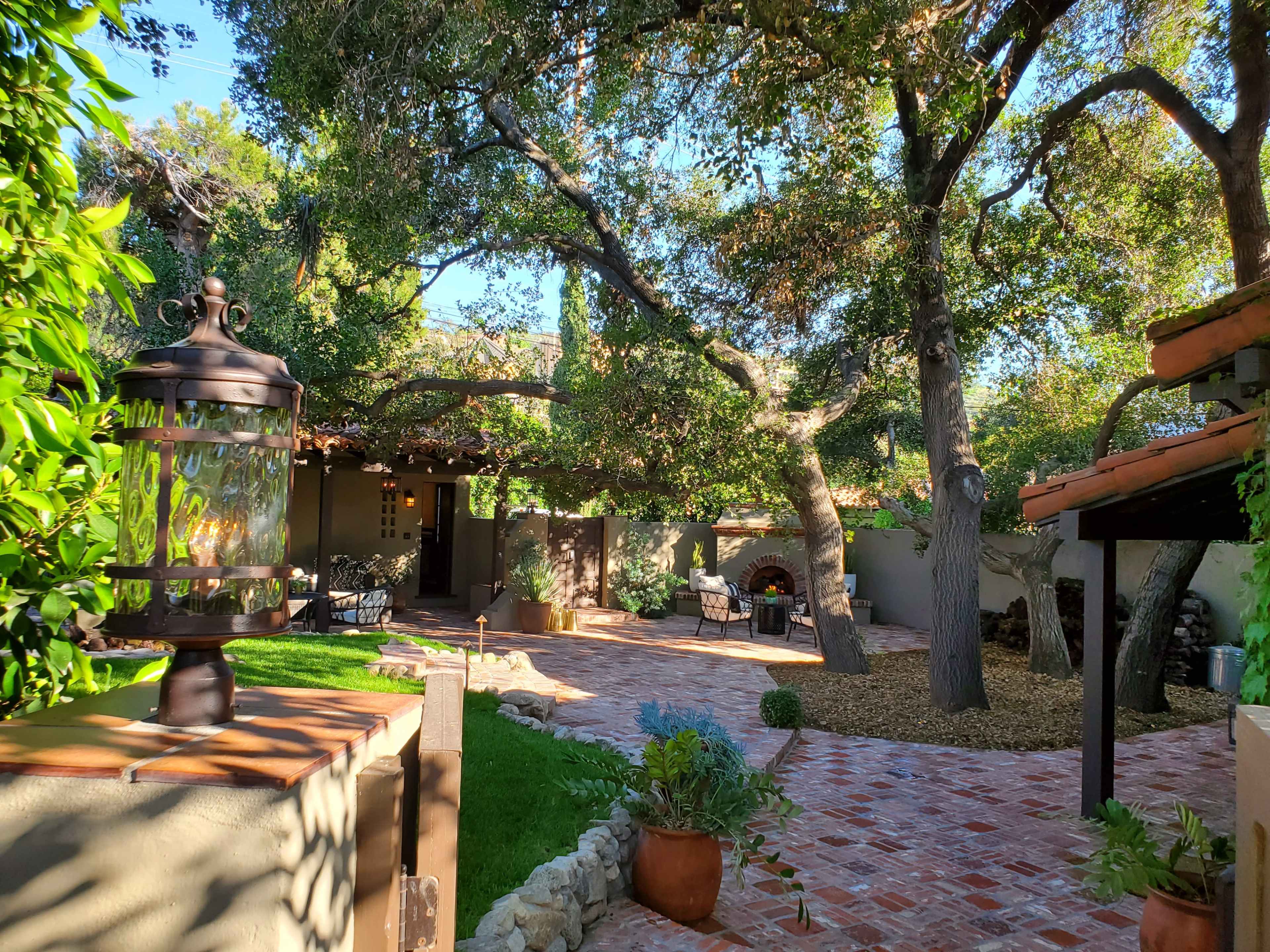 The image shows a landscaped garden area with stone pathways, seating arrangements, and large trees providing shade.