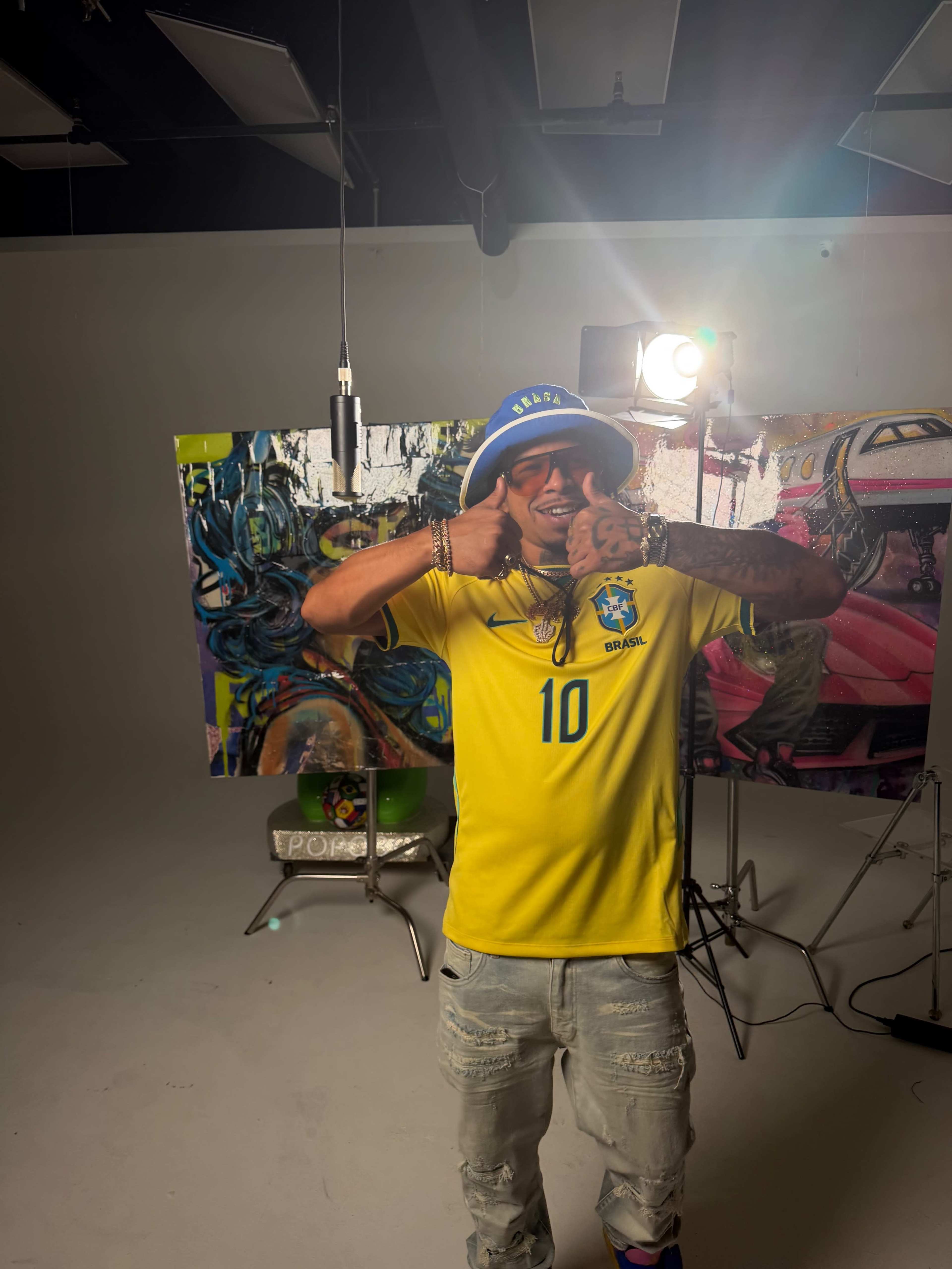 A person wearing a yellow Brazil soccer jersey and a blue hat poses playfully in a studio with colorful artwork in the background.