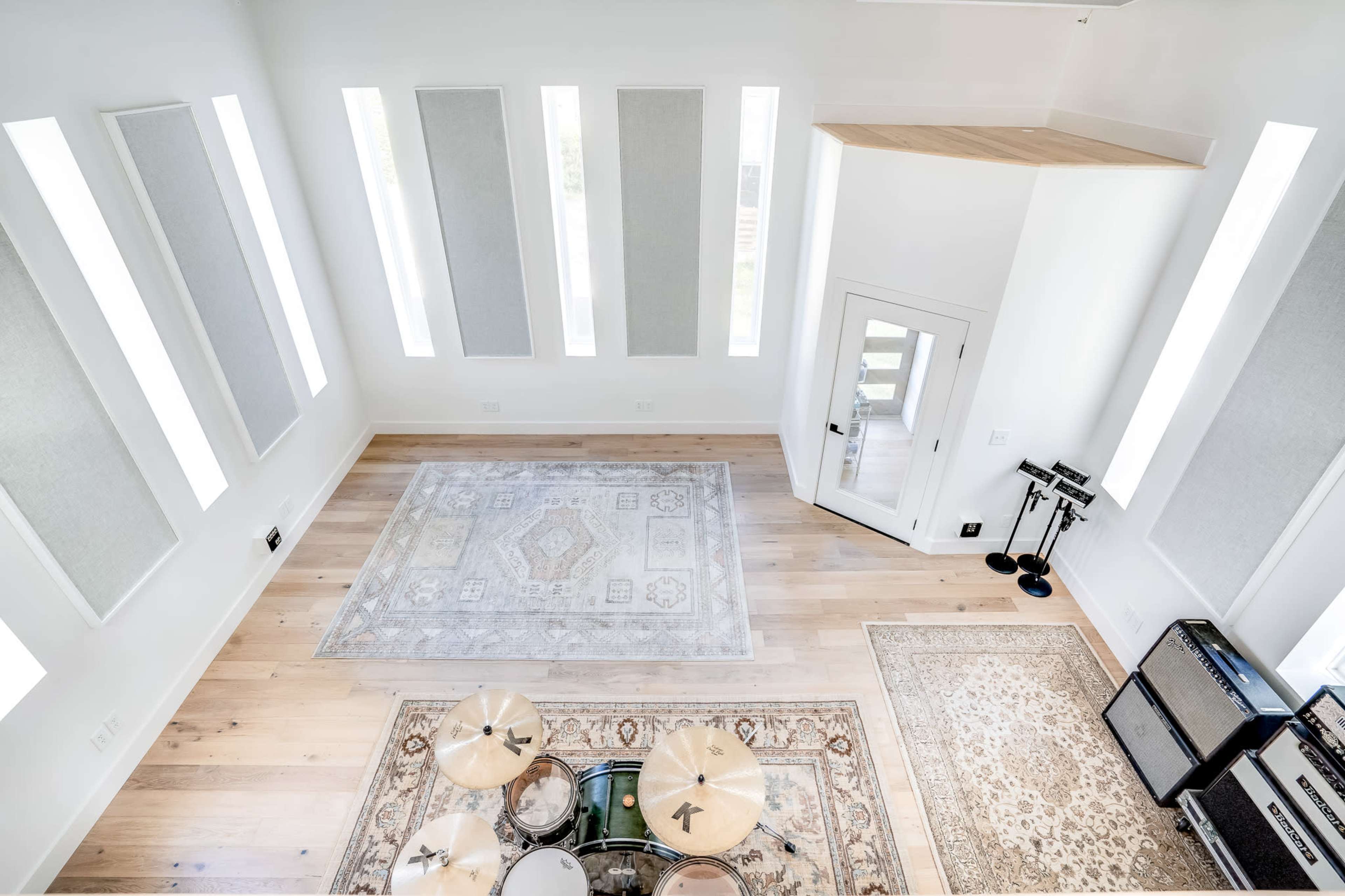 A spacious, well-lit room with wooden floors, two area rugs, and a drum set in one corner, along with an amplifier setup nearby.