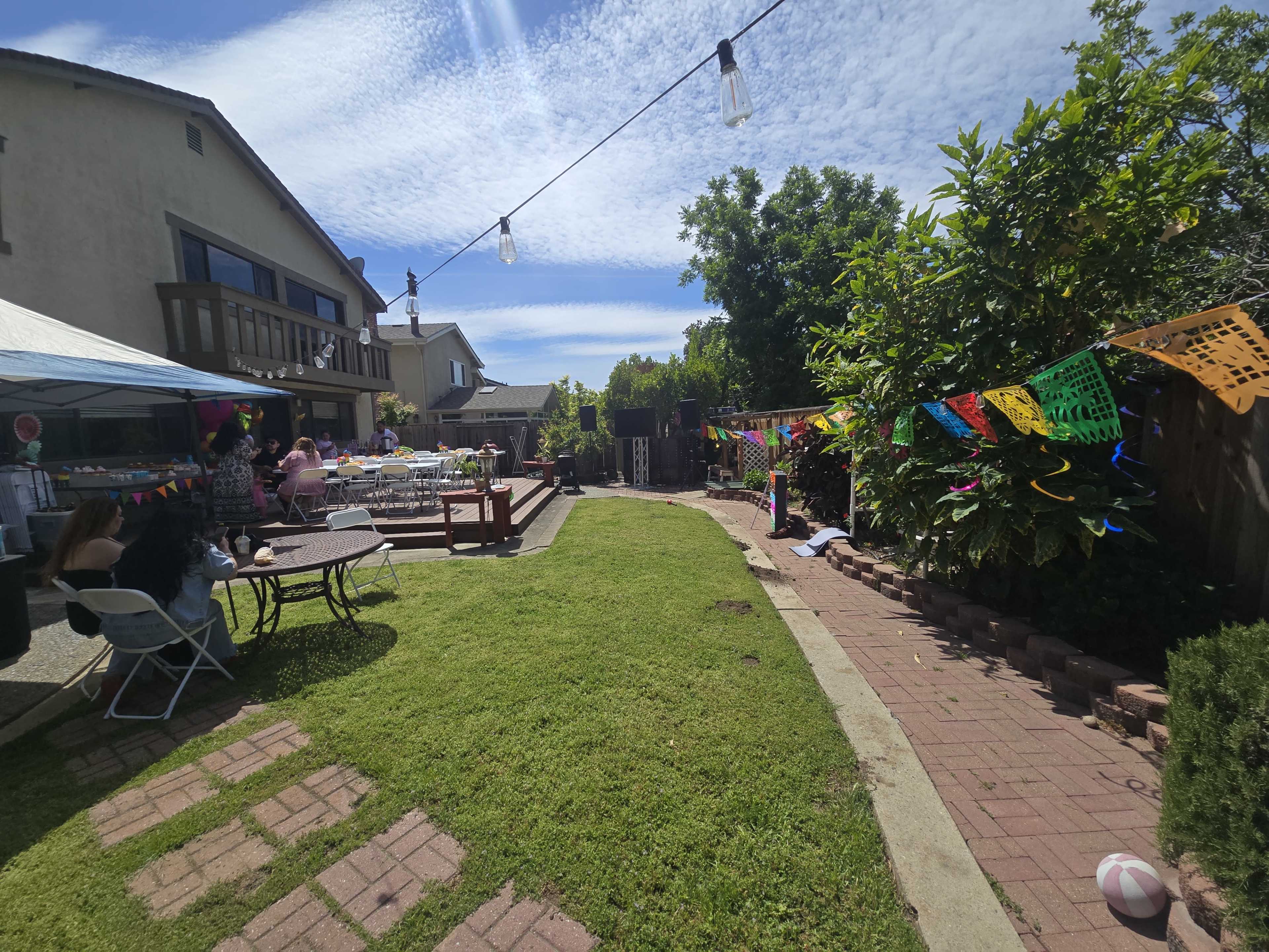 The image depicts a sunny backyard party with colorful decorations, tables set for guests, and a well-manicured lawn.