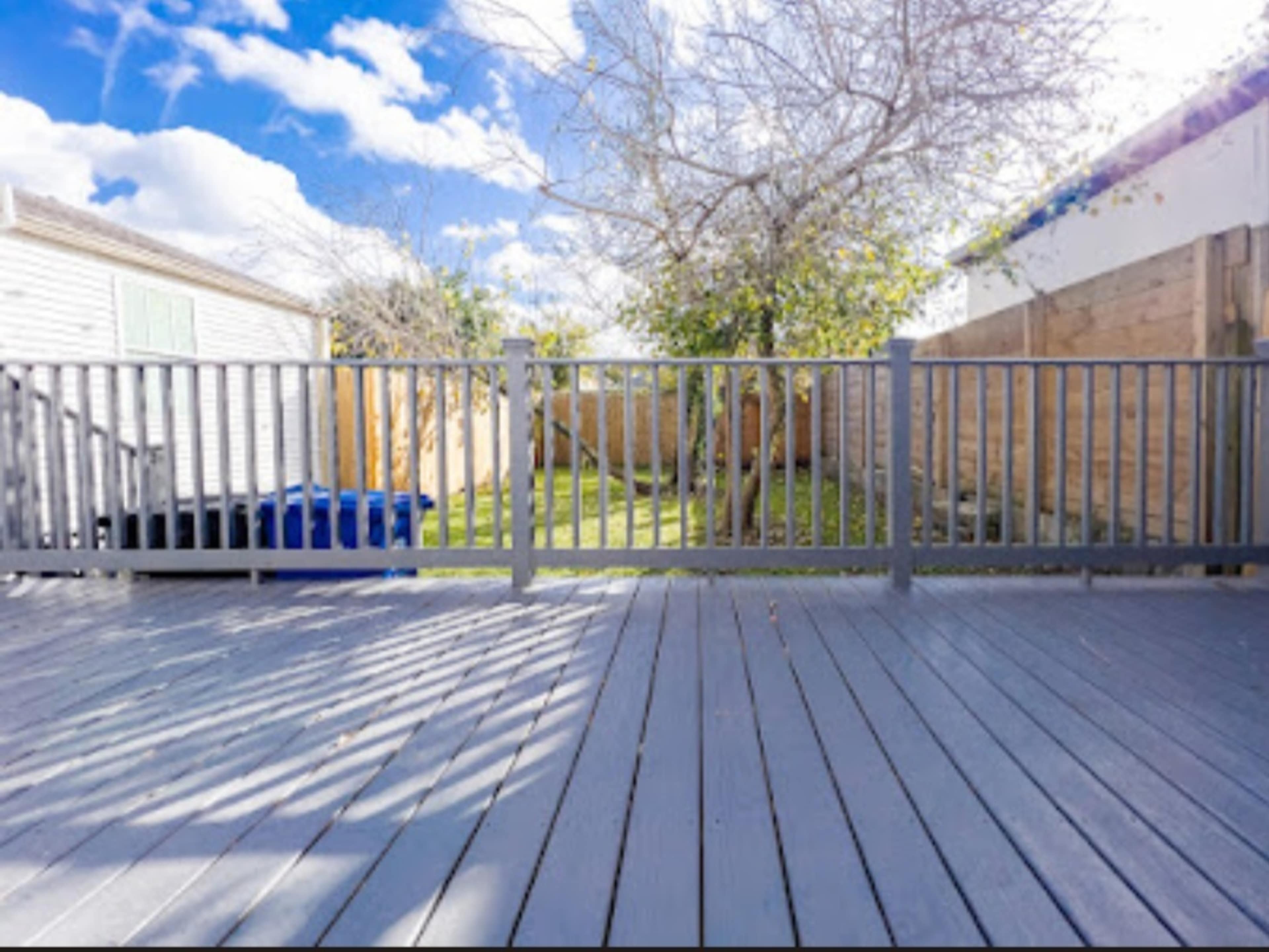 A wooden deck with a gray finish overlooks a grassy backyard surrounded by a wooden fence and several trees.