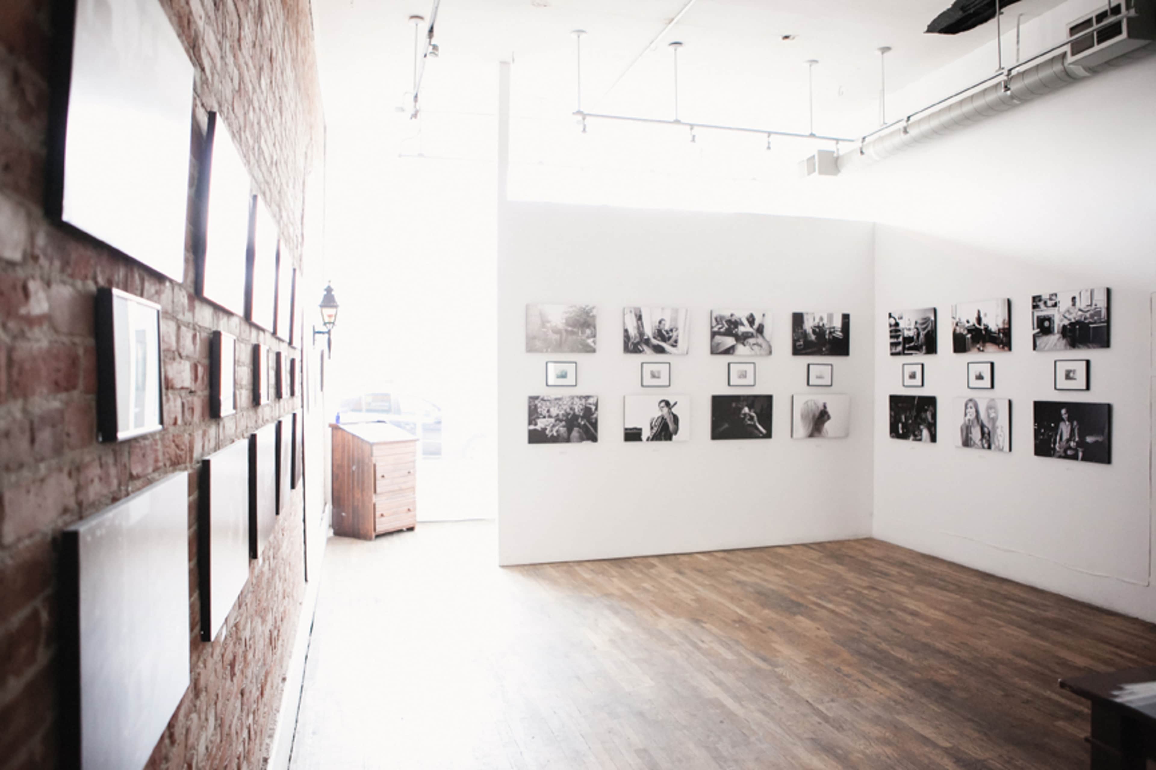 An art gallery with a brick wall on the left displaying black and white photographs, and a white wall on the right featuring more framed photographs.